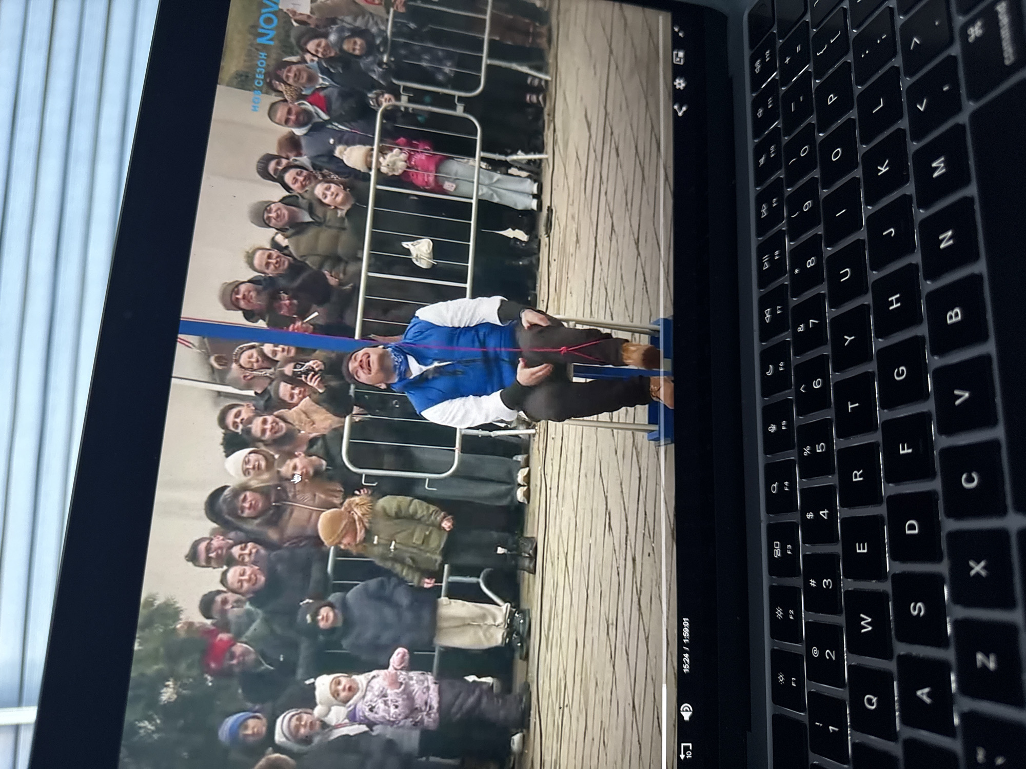 The image shows a laptop screen displaying a video of a crowd of people behind metal barricades. A man wearing a blue and white shirt is being supported by the barricades. The setting appears to be outdoors, with a light-colored paved surface visible. The video is likely in playback mode based on the timestamp at the bottom. A portion of the laptop's keyboard is also in the frame, with the keys backlit.