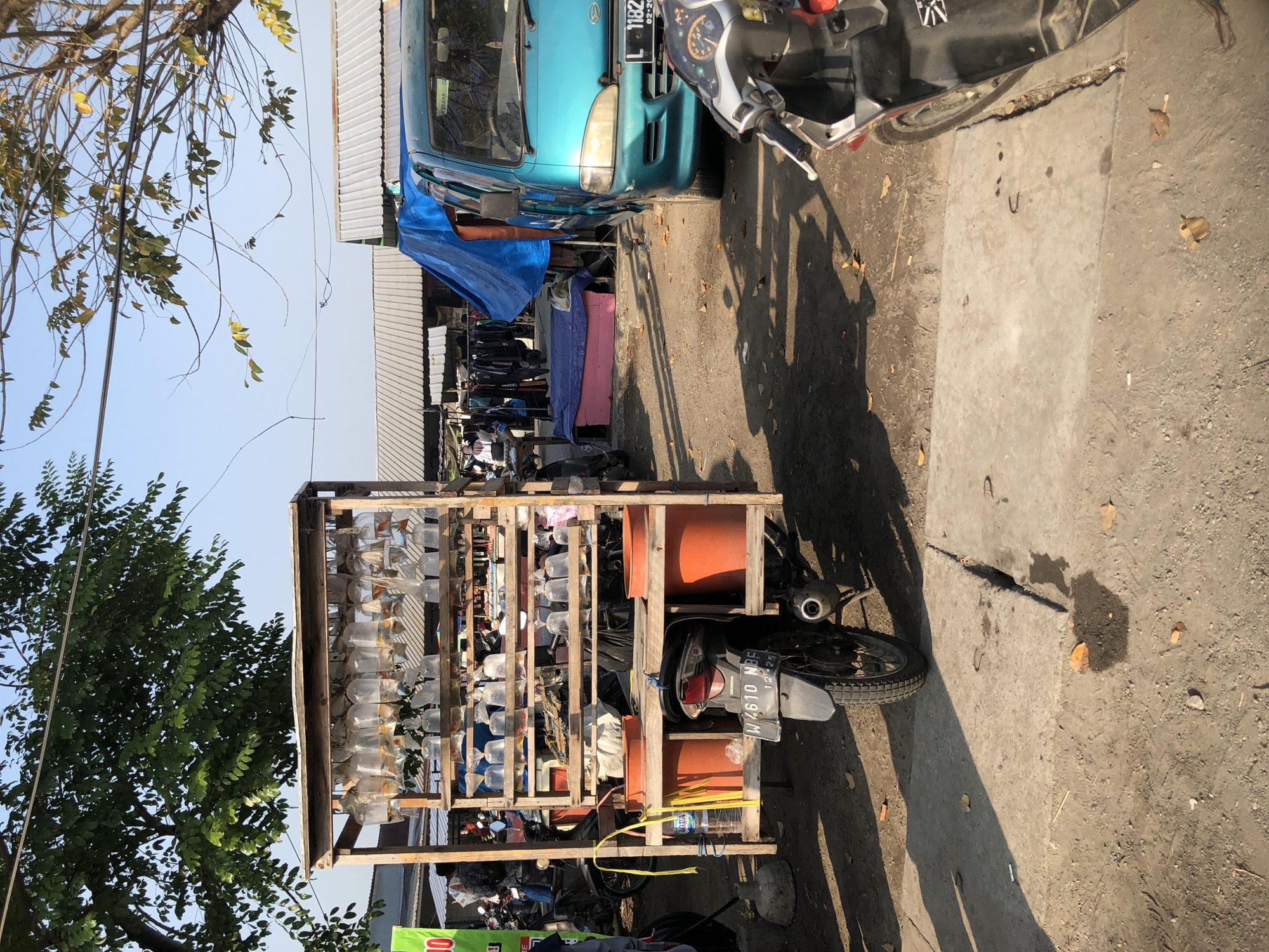 An outdoor market scene in Balongdowo, Indonesia, features a wooden cart loaded with clear plastic bags of fish, suspended from a rack. The cart is attached to the back of a motorcycle. The ground is a mixture of dirt and concrete. In the background, various stalls with corrugated metal roofs are visible, along with a bright blue tarp covering some items and a turquoise van. A scooter is parked in the foreground, partially obscuring the view of the motorcycle. The sky is clear and blue, and trees with green and yellowing leaves cast shadows on the ground. Several people are present in the background, some tending to market stalls. The overall impression is of a bustling, informal marketplace.