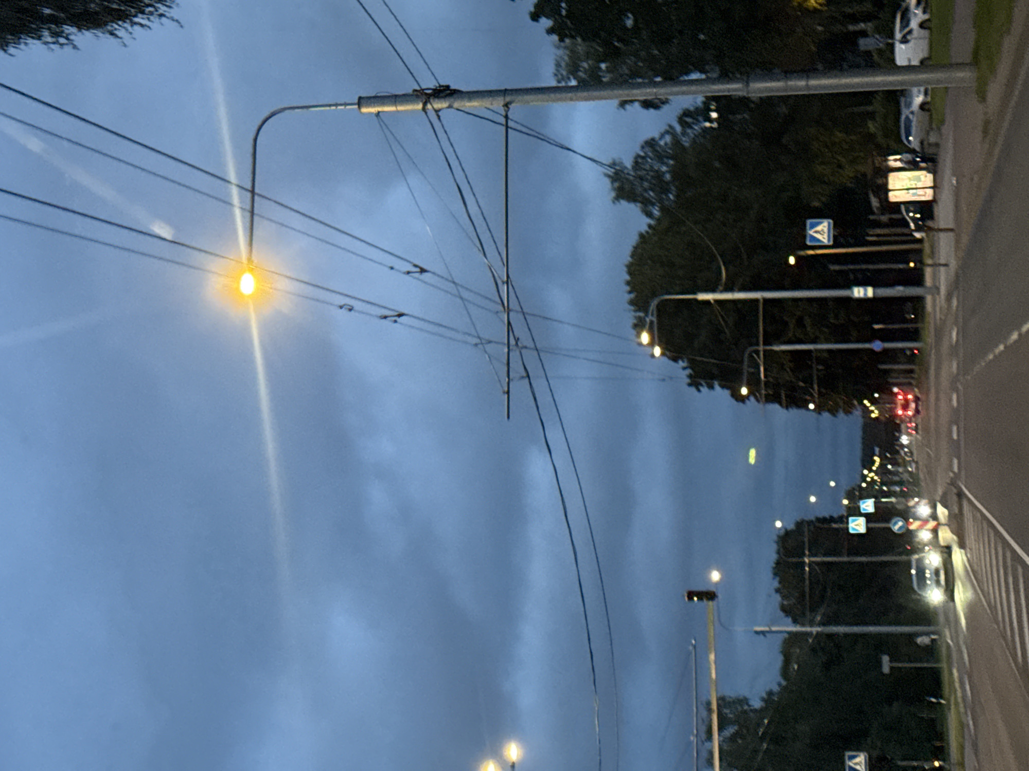The image depicts a street scene in Tallinn, Estonia, during dusk or early night. The sky is overcast with dark, moody clouds, and streetlights are illuminated, casting a warm glow. A prominent streetlight with a bright yellow lamp is visible on the right side of the frame, supported by a tall pole. Overhead, a complex network of trolleybus wires crisscrosses the sky. The street itself is paved and appears to be a multi-lane road with traffic. Several cars are visible, some with their headlights on, including a white car in the foreground approaching a pedestrian crossing. Pedestrian crossing signs, with blue backgrounds and white pedestrian symbols, are positioned at intervals along the roadside. Trees line the sides of the street, and in the distance, more streetlights and building lights can be seen, suggesting an urban or suburban environment. The overall atmosphere is calm and captures the transition from day to night in a city setting.