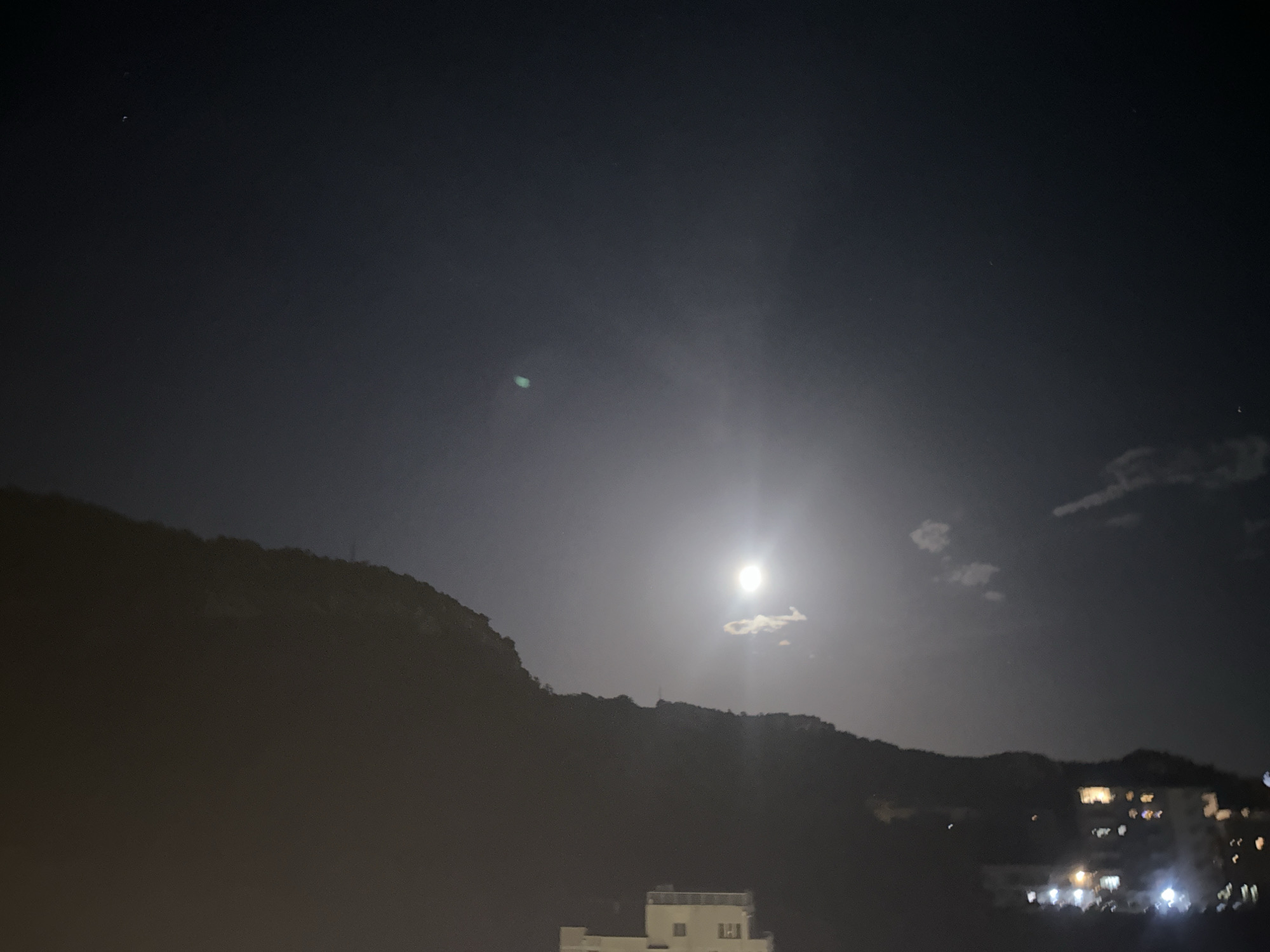 This is a nighttime photograph taken from a slightly elevated position in Rafailovići, Montenegro. The scene is dominated by a dark, mountainous silhouette on the left, with the full moon shining brightly in the sky, slightly to the right of the center. A few small, wispy clouds are visible near the moon, and there are lights from buildings and streetlights on the right side of the image, indicating a populated area. The sky is dark blue, with a few faint stars visible. The foreground shows the tops of some buildings, with one white, modern-looking structure prominently displayed at the bottom. The overall atmosphere is serene and typical of a clear night in a coastal town.