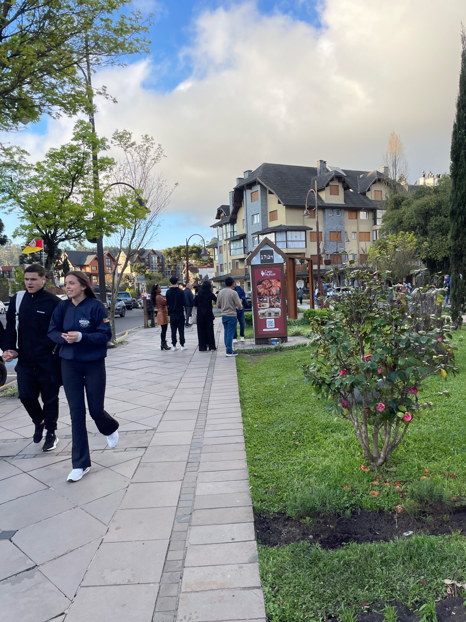 The image shows a street scene in Gramado, Brazil, with a sidewalk lined with pavers and a grassy area. Several people are walking along the sidewalk. On the left, a young couple is walking, with the male in a black tracksuit and the female in a blue jacket and black pants. Further down the sidewalk, a group of people are walking, including individuals in dark clothing and one person in a light-colored top and jeans. The background features buildings with pitched roofs, characteristic of European-influenced architecture, and trees. A tall evergreen tree stands on the right side of the frame. A sign with an advertisement for "Casa Muttoni" is visible near the center of the image, and a flowering bush with pink blossoms is in the foreground on the right. The sky is partly cloudy.