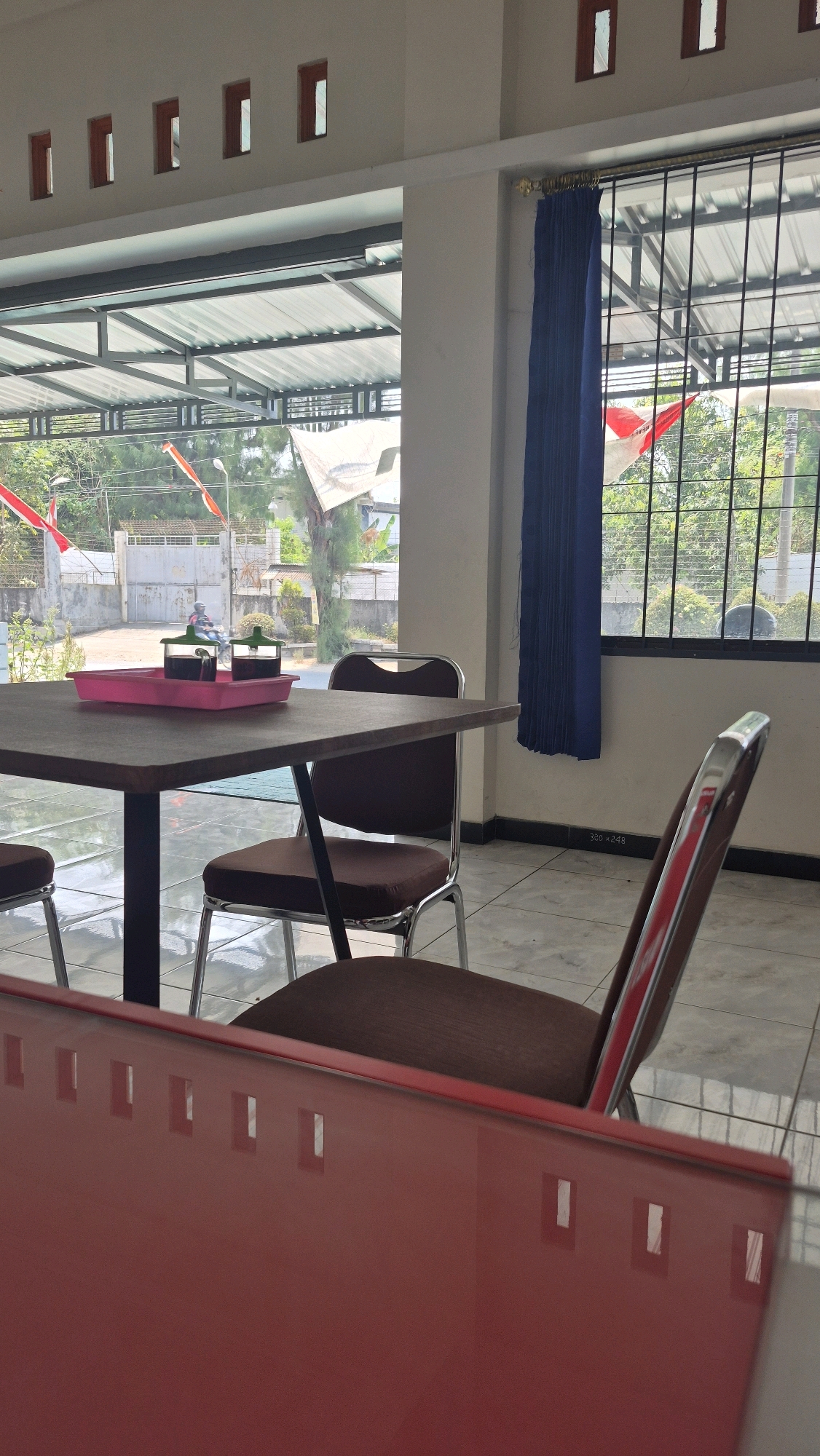 A casual dining area in Bojonegoro, Indonesia, features a square table made of dark wood with a metal frame. Two chairs with brown upholstery and metal frames are positioned at the table. A pink tray holds two small dark bottles, presumably condiments. The room has white walls and a tiled floor. Large windows with metal bars look out onto a street with trees and buildings, and a red and white flag is visible. A blue curtain hangs to the right of the window. In the foreground, a portion of a red table or counter with a reflective surface is visible. The scene appears to be an outdoor or semi-outdoor dining space, possibly at a restaurant or cafe.