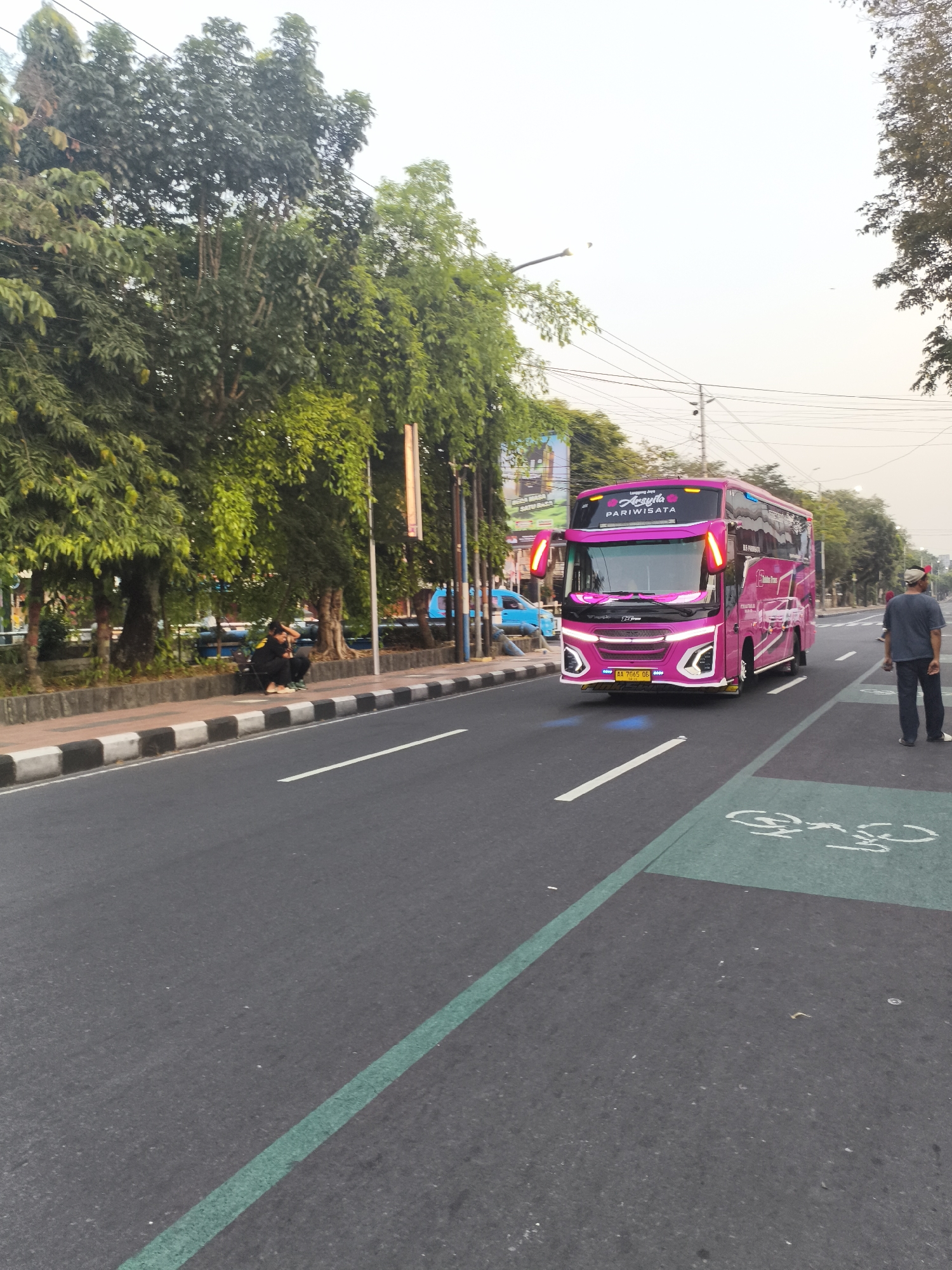 A bright pink double-decker tour bus drives on a city street in Magelang, Indonesia. The bus has "Asyila PARIWISATA" written on its front. A man in a dark shirt and pants stands on the right side of the street, facing away from the camera. Two people sit on a sidewalk to the left of the street, near a line of trees. The street is paved and has a green bike lane marked on the right. The sky is overcast.