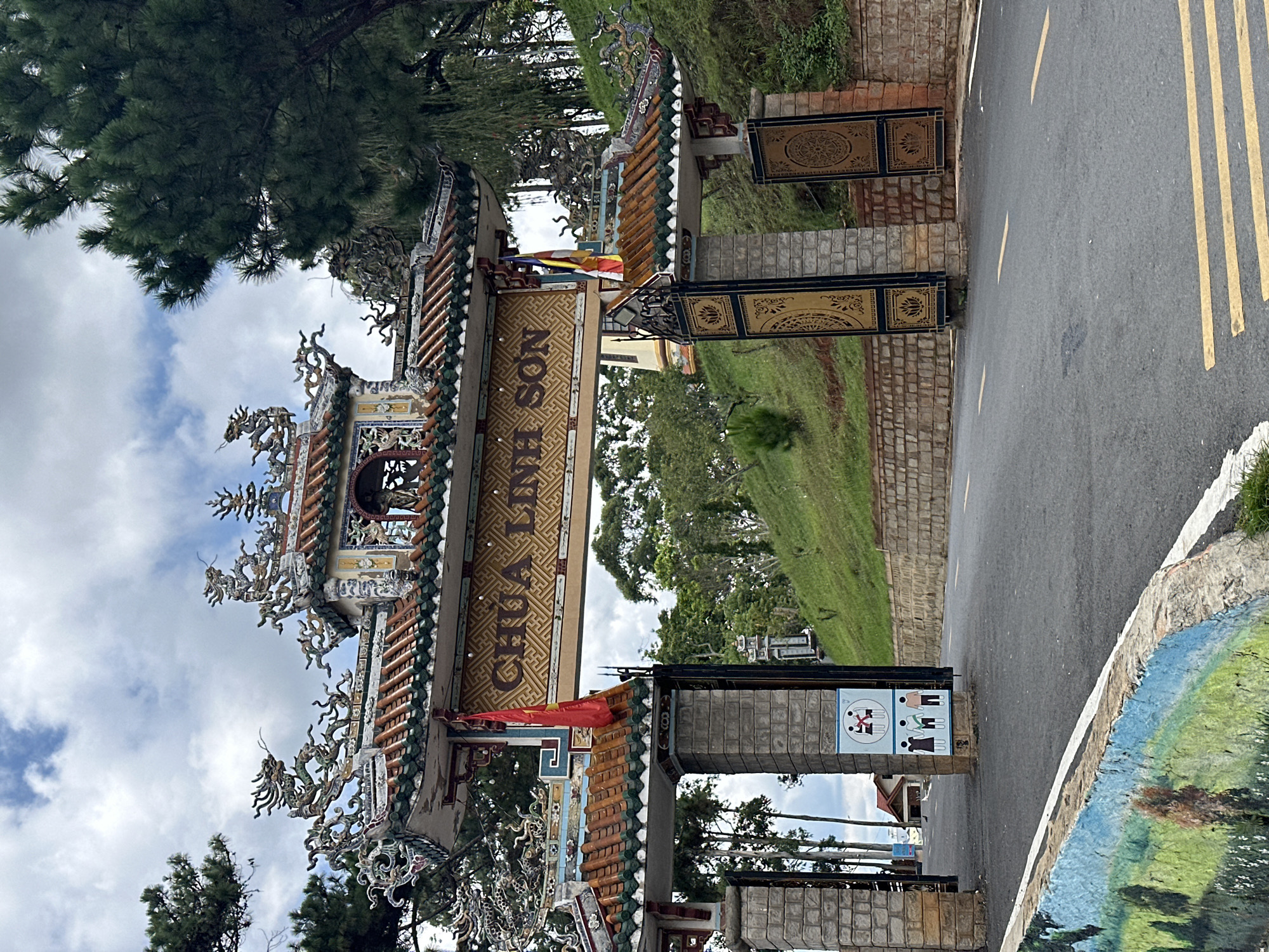 The image shows the entrance gate to Linh Son Pagoda in Da Lat, Vietnam. The gate is an elaborate structure with traditional Vietnamese architecture, featuring ornate roof decorations, dragon motifs, and a large sign with Vietnamese characters. The gate is made of stone pillars and ornate metalwork. A red Vietnamese flag and a Buddhist flag are hung from the structure. The entrance leads to a paved road that curves uphill, with lush green hills and trees on either side. In the foreground, a low wall with a colorful mural is visible. The sky is cloudy with patches of blue. There are no people visible in the image.