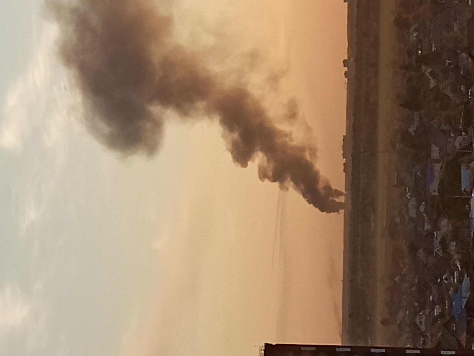The image shows a large plume of dark smoke rising into the sky over a city, likely Кондратово, Russia, based on the context. The scene is shot from a relatively high vantage point, possibly from a building overlooking a residential area. The sky is hazy with a warm, golden hue. Buildings and houses are visible in the foreground and middle ground, suggesting an urban setting. The smoke plume is the dominant feature, indicating a fire or explosion of considerable magnitude.