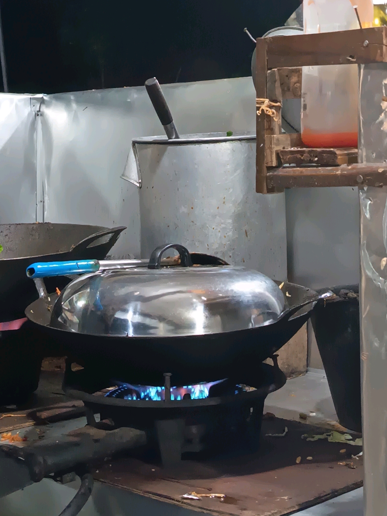 The image captures a close-up view of a wok cooking station, likely at an outdoor food stall in Rantopanjang, Indonesia. A large, dark wok sits atop a gas burner with visible blue flames igniting the cooking surface. The wok is covered by a shiny, dome-shaped lid, with a bright blue handle protruding from one side. To the left of the wok, another dark pot or wok is partially visible, and a utensil with a dark handle rests on the rim of a large metal container in the background. Above and to the right, a rudimentary wooden frame holds a clear plastic container filled with an orange liquid. The surrounding environment appears to be part of a makeshift kitchen setup, with metal sheeting forming the backdrop and a worn countertop in the foreground. The lighting suggests it is evening or nighttime, with artificial light sources illuminating the scene.
