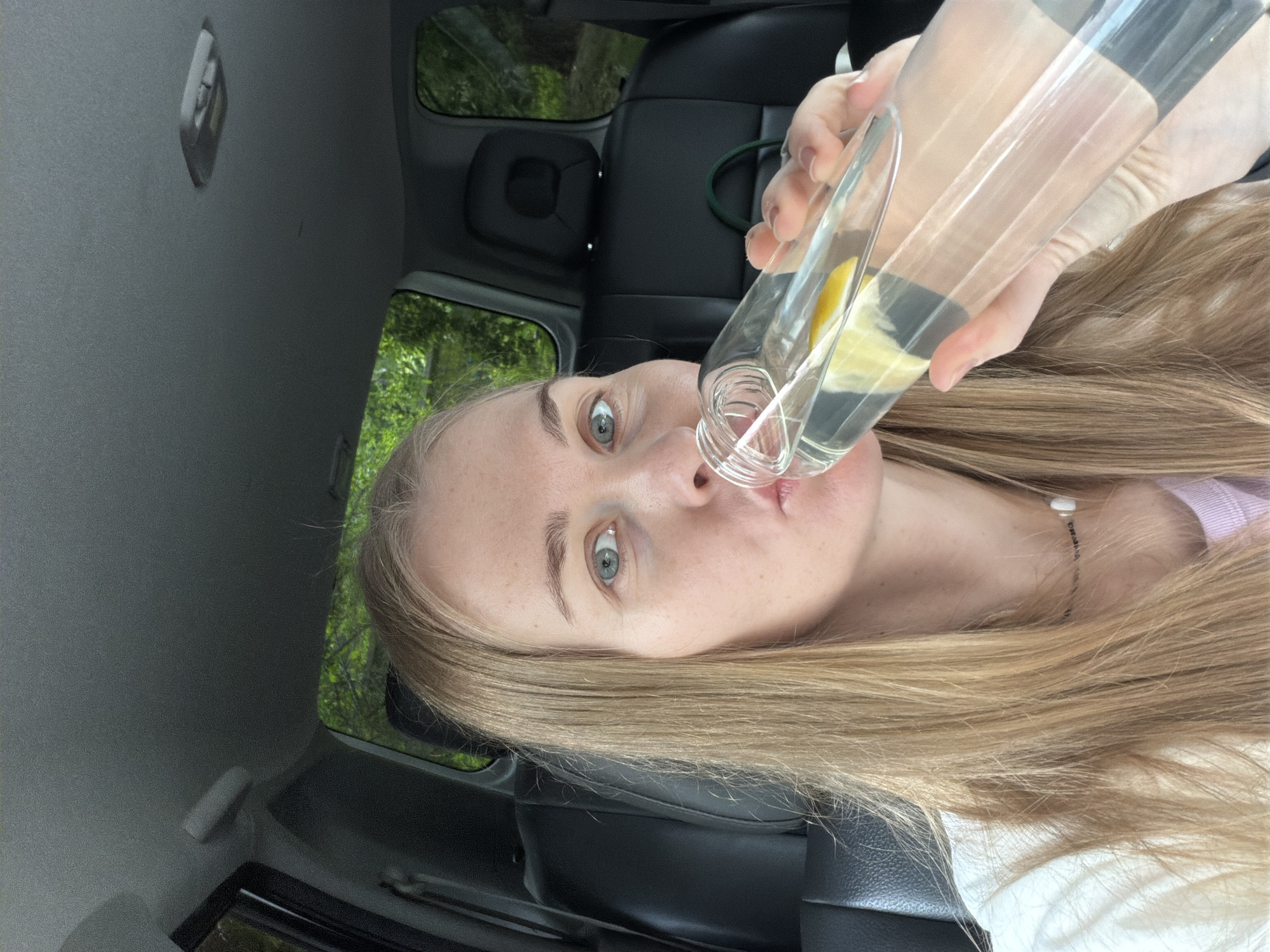 A blonde woman with blue eyes is sitting in the passenger seat of a car and drinking from a clear glass bottle of water with lemon slices. The interior of the car is visible, including black leather seats and the car's headliner. Outside the car windows, lush green trees are visible, suggesting a natural or suburban setting. The woman is wearing a white t-shirt and a light pink top underneath, with a necklace around her neck. The image is a selfie taken from a slightly low angle, focusing on the woman's face and the bottle.