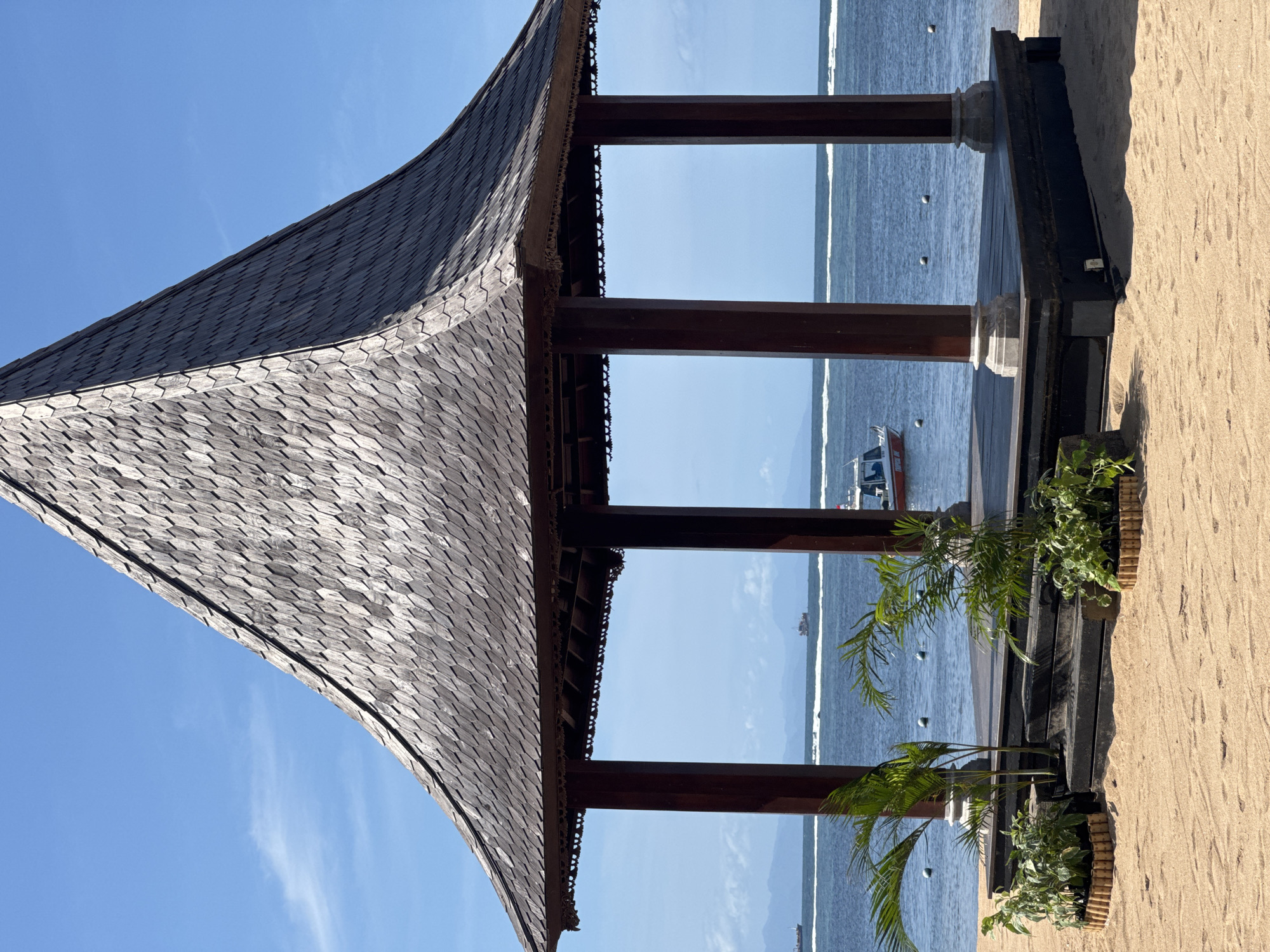 The image depicts a pavilion or gazebo located on a beach in Kuta Selatan, Indonesia. The structure has a distinctive roof covered with small, overlapping tiles, possibly made of wood or a similar material. The roof curves gracefully and overhangs the supporting beams. The pavilion is open-sided, with dark wooden columns providing structural support and framing the view of the ocean. The columns are connected by horizontal beams at the top and bottom.
The beach appears to be sandy, with the edge of the frame showing the texture of the sand. In the background, the ocean is visible, with boats or other watercraft dotting the horizon. The sky is clear and blue, suggesting a sunny day. The gazebo sits atop a dark wooden platform, with vegetation, like small palm trees and other plants, sitting in decorative planters on the platform's edge.