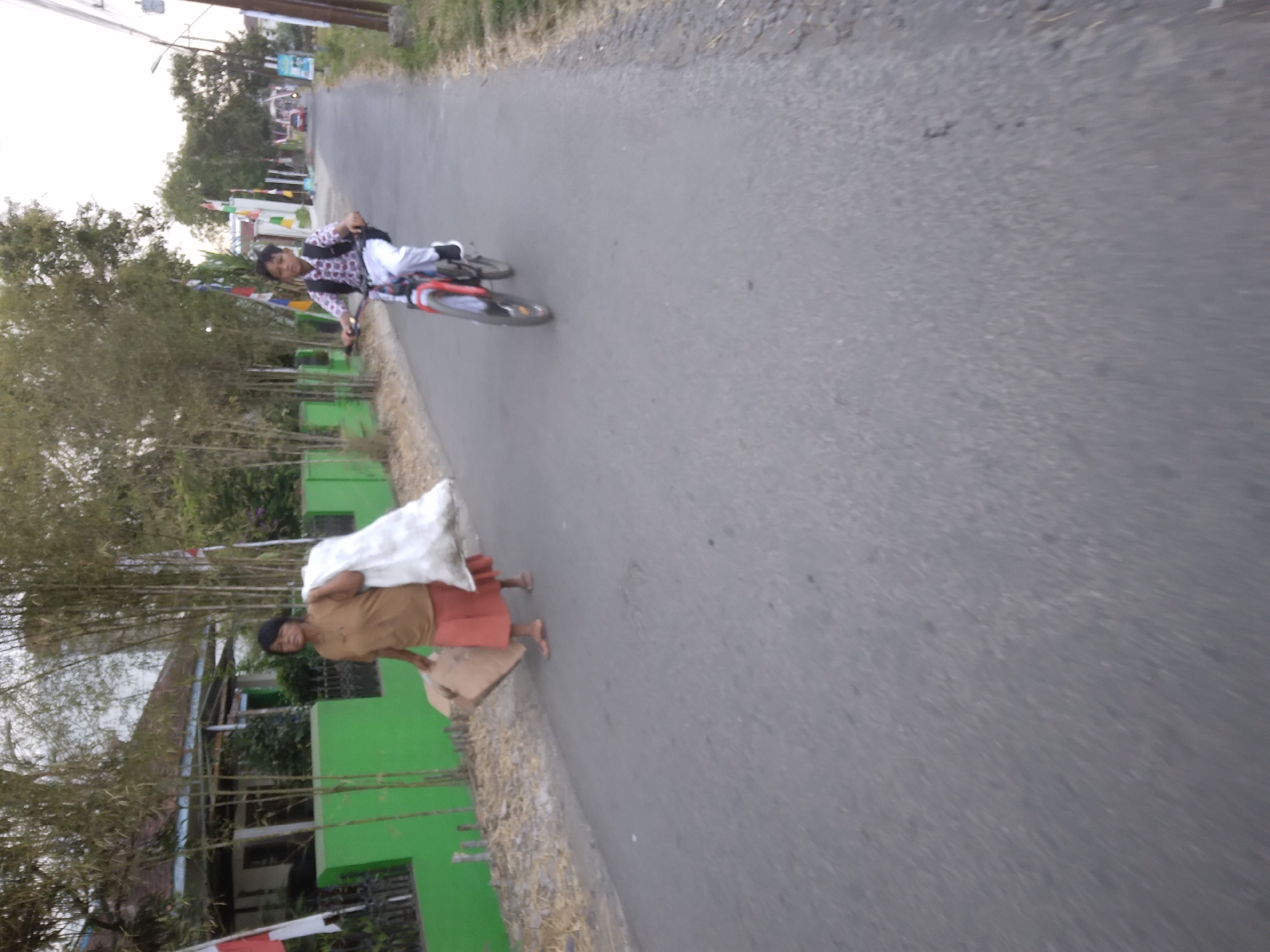 In Desa Pamijen, Kecamatan Sokaraja, Indonesia, an outdoor scene unfolds on an asphalt road. A woman, wearing a light brown top and an orange skirt, walks along the side of the road carrying a large white plastic bag over her left shoulder and a cardboard box in her right hand. Behind her, a young boy rides a bicycle with a red frame, wearing a patterned shirt and white pants, with a black backpack on his shoulders. The road is bordered by a green fence and bamboo trees, with a building visible behind the fence. The sky is overcast, suggesting either early morning or late afternoon.