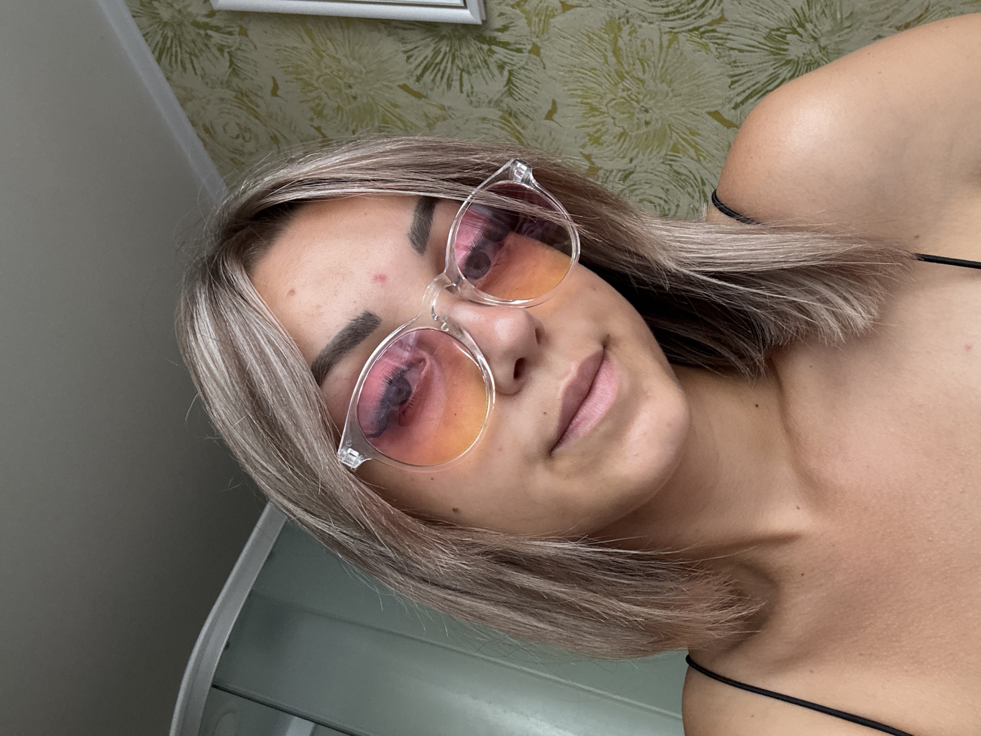 The image shows a close-up, head-and-shoulders shot of a young woman with blonde hair, wearing clear-framed sunglasses with gradient pink and orange lenses. She has makeup on, including mascara and defined eyebrows, and a small blemish on her forehead. Her shoulders and upper chest are visible, and she is wearing a black spaghetti strap top. The background includes a portion of a wall with floral wallpaper and a sliver of a light-colored shower or cabinet. The lighting suggests an indoor setting, likely a bathroom or bedroom. The photo appears to be a selfie. There is no visual information within the image to determine the location within Krasnogorskoye, Russia.