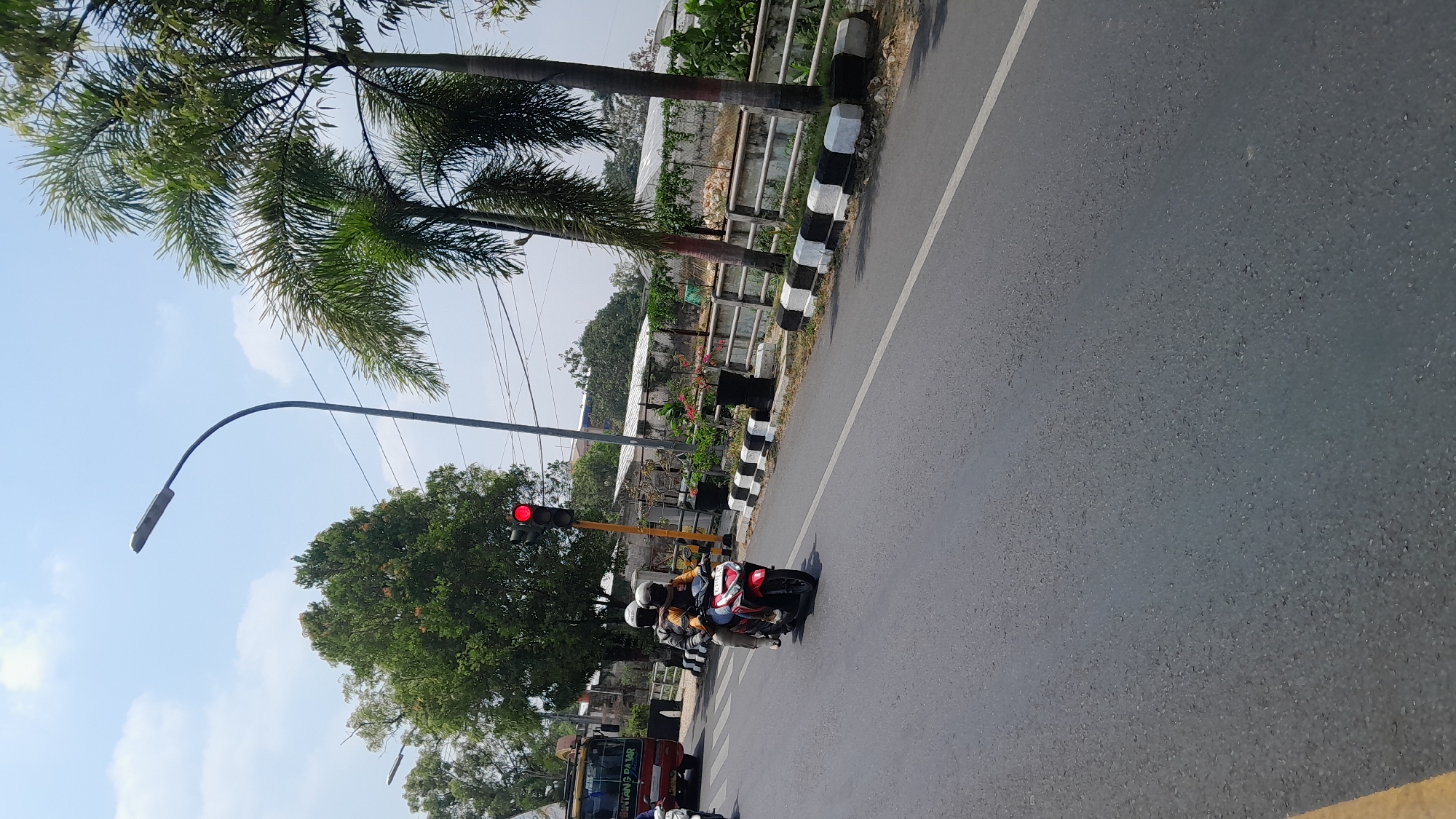 The image depicts a street scene in Blora, Indonesia, on a clear, sunny day. In the foreground, a red scooter with two occupants wearing helmets is traveling on an asphalt road. The scooter has a license plate that is partially obscured. Behind the scooter, a red bus with "BINTANG PANJAR" written on its side is visible, also traveling on the road. A traffic light is illuminated red, positioned on a yellow pole. To the right of the road, a white fence with black and white striped concrete barriers runs along the side. Several palm trees and other greenery are present. The sky is blue with scattered white clouds. The scene appears to be a typical urban or suburban street with moderate traffic.