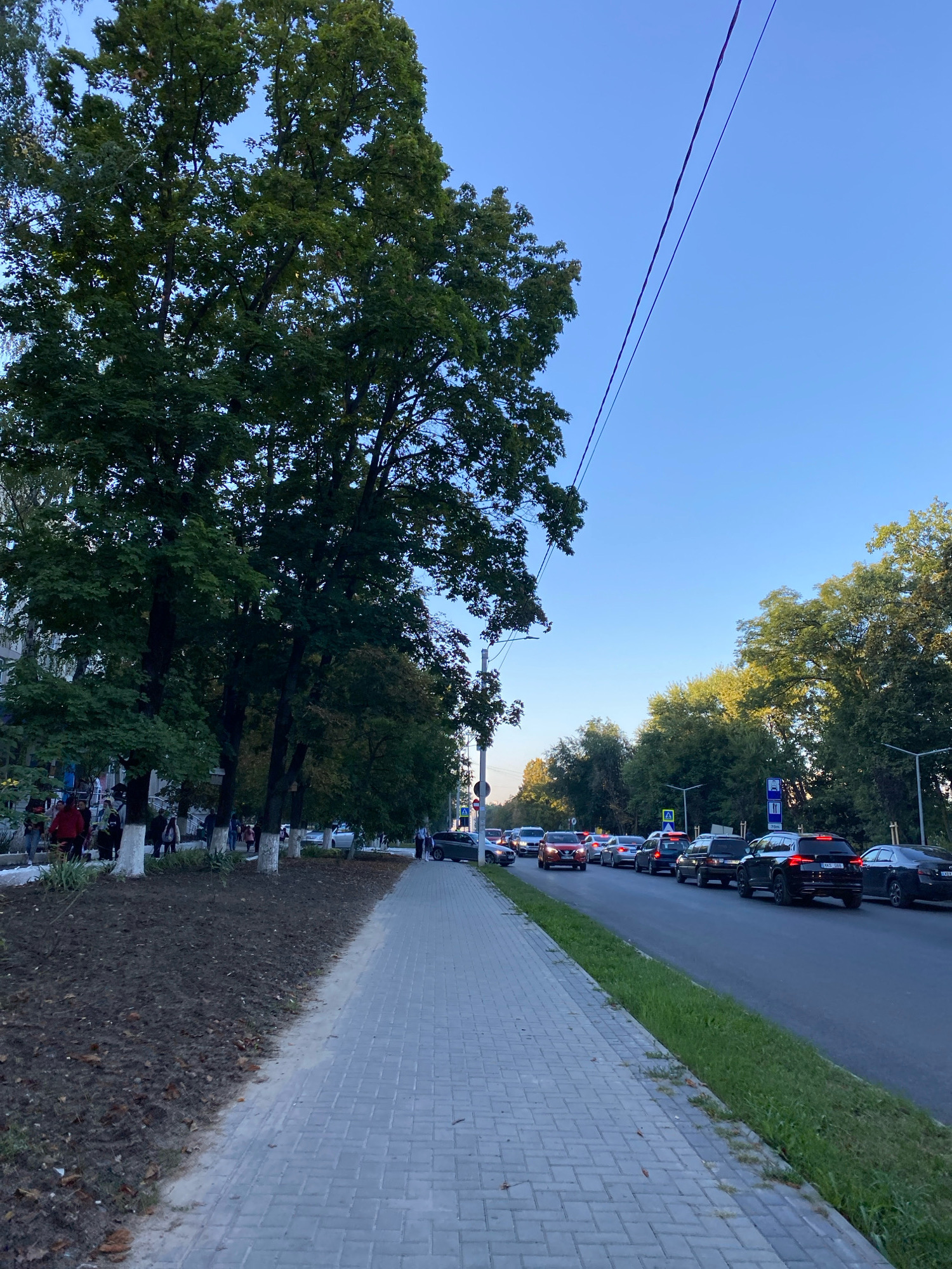 A sidewalk runs alongside a street filled with cars in Chișinău, Moldova. On the left side of the sidewalk, there are trees with green leaves and a building. People are walking on the sidewalk. On the right side of the street, there are more trees and a clear blue sky. A power line is visible stretching across the sky. The scene depicts a typical urban environment with traffic and pedestrians.