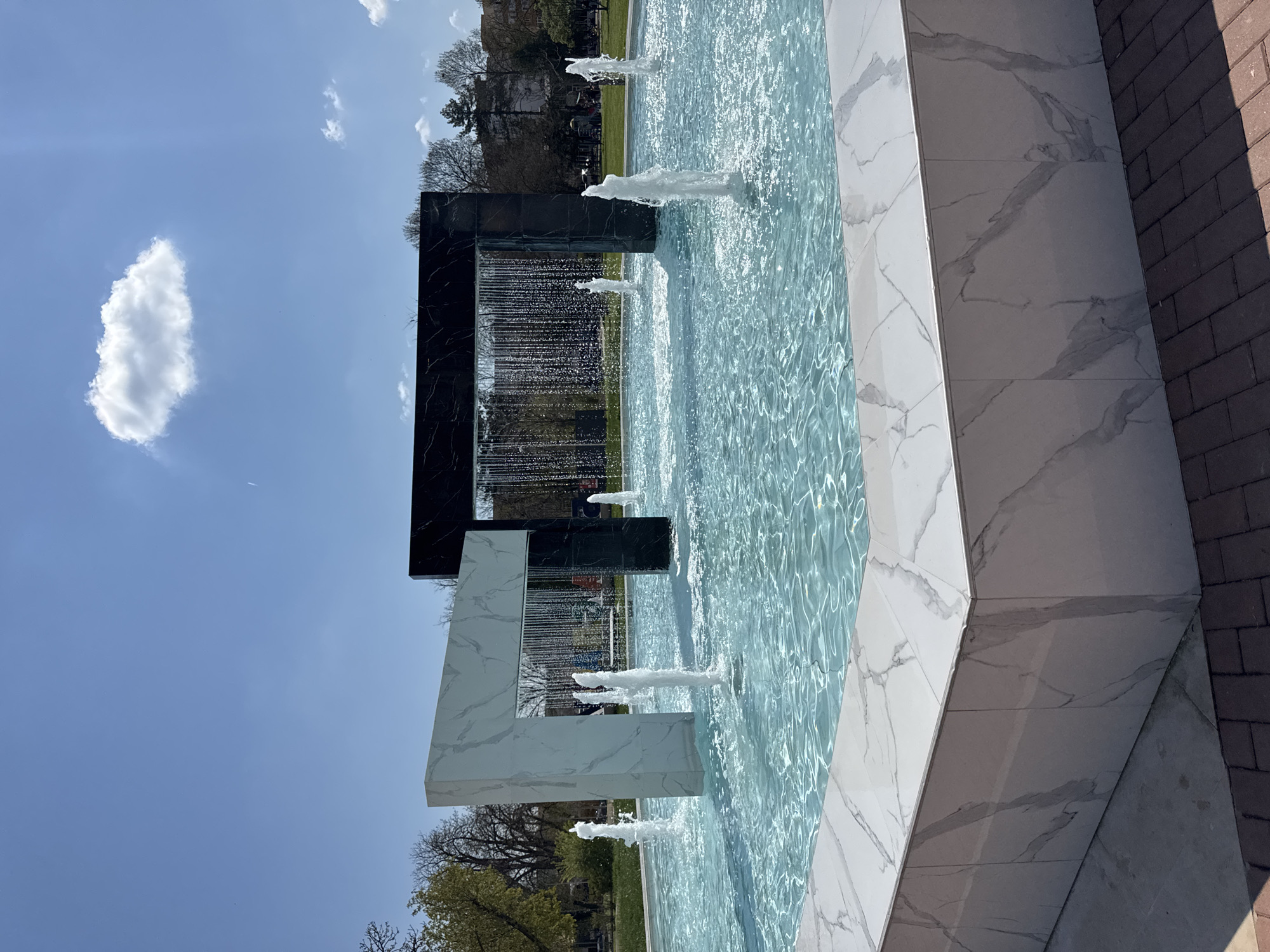 A photograph captures a rectangular fountain with a white marble exterior, filled with clear water, and featuring multiple water jets. The fountain's design incorporates dark-colored slabs forming an archway-like structure within it. The surrounding environment includes green grass, trees, and distant buildings. A blue sky with a single, prominent white cloud is visible overhead. The photograph appears to be taken in daylight and from a slightly elevated perspective. The location is Byala Slatina, Bulgaria.