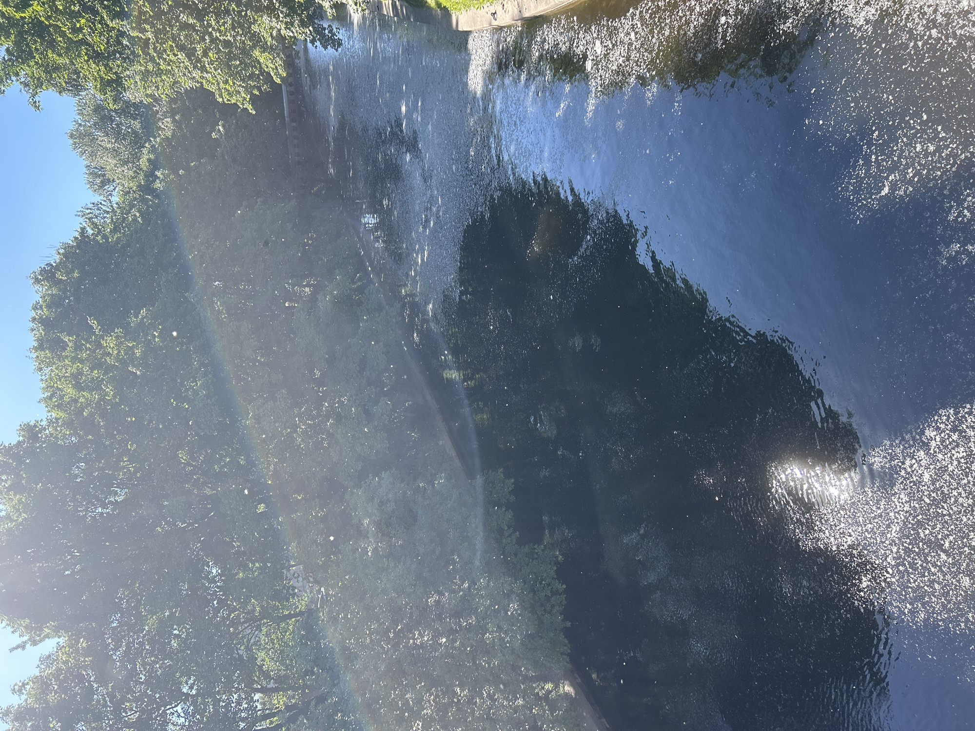 The image displays a reflective body of water, likely a canal or river, bordered by lush green trees. The water's surface is disturbed by ripples, causing the reflections of the trees and the sky to appear distorted. Sunlight glints off the water, creating a shimmering effect in some areas, while other parts are darker, reflecting the shadows of the surrounding foliage. A faint rainbow arc is visible in the upper left portion of the image, suggesting the presence of mist or spray in the air, possibly from a fountain or waterfall. The sky is a clear, bright blue. The scene is bathed in natural light, indicating it is daytime, likely on a sunny day given the bright reflections on the water. The overall impression is one of a tranquil natural setting, possibly a park or a quiet waterway. There is no visible text in the image.
