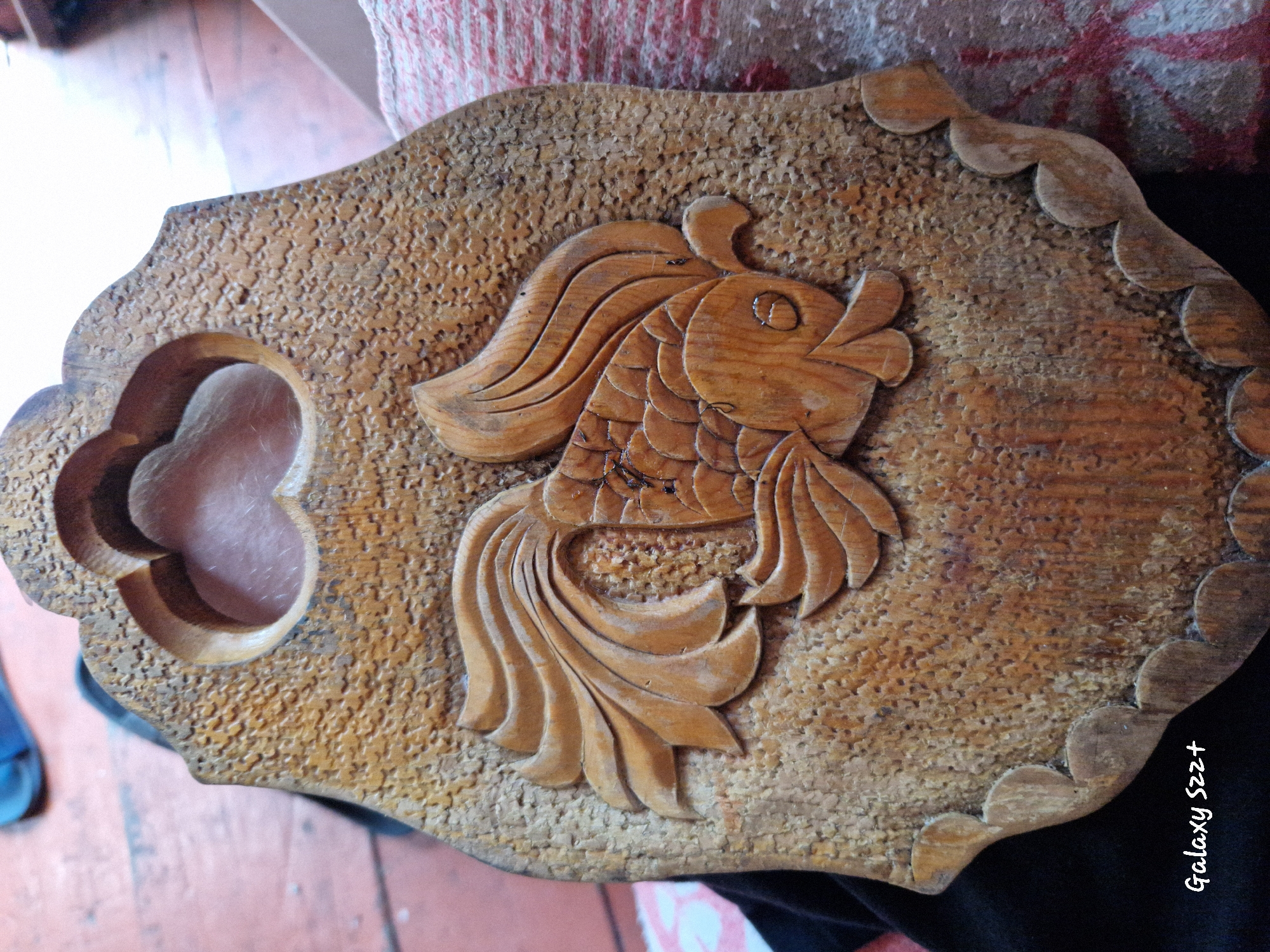 The image shows a close-up, top-down view of a wooden carving of a goldfish. The carving is intricate, with detailed scales, fins, and a decorative, scalloped border along the bottom edge. The wood appears to be aged and has a warm, brown tone with visible grain. The background is out of focus, but suggests a wooden floor and possibly some fabric. The bottom left corner of the image contains the text "Galaxy S22+". There is no information in the image to suggest its location in the city of Myski, Russia.