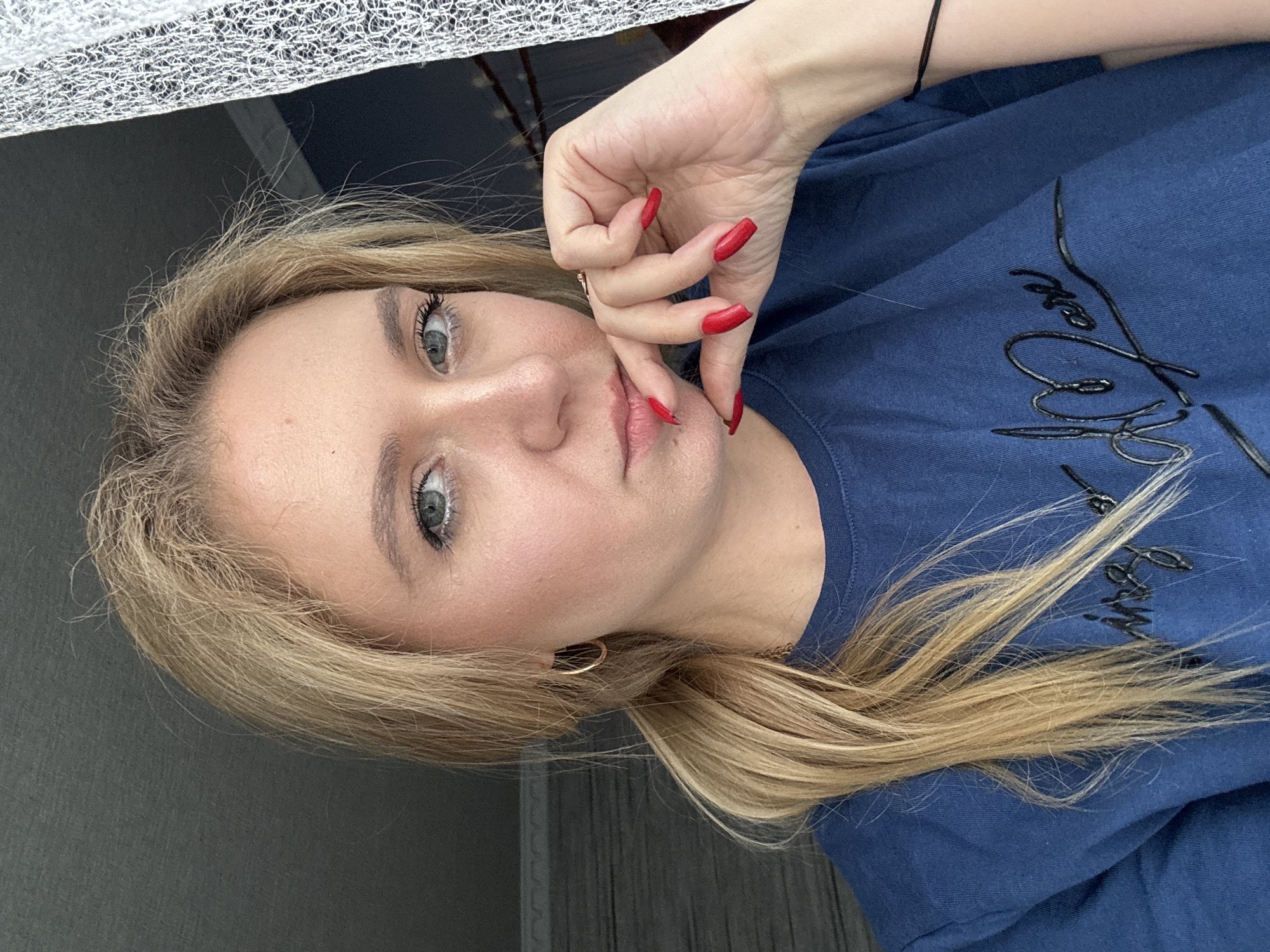 A young woman with blonde hair tied in a ponytail, wearing a navy blue t-shirt, is shown in a close-up shot. She has her hand resting on her chin, with bright red painted fingernails. Her eyes are green and she is looking directly at the camera. The background appears to be a room with a dark wall on the left and a white, sheer curtain on the right. The lighting suggests it is daytime, possibly near a window.