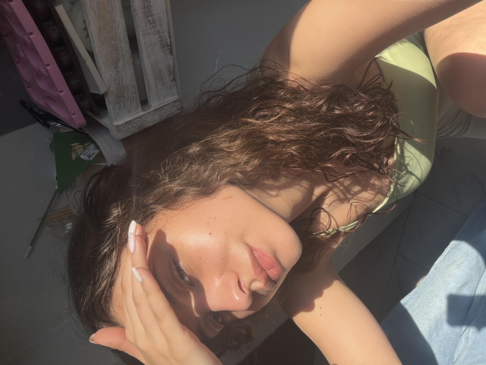 The image captures a young woman lying down, viewed from a high angle, with strong natural sunlight illuminating her. She has long, wavy, dark brown hair and is wearing a light green top or swimsuit. Her right hand is positioned near her forehead, displaying white or iridescent nail polish. Her face, including her nose and lips, is well-lit, highlighting her natural skin texture, while parts of her face and body are in shadow. Her expression is neutral and relaxed as she looks towards the camera.
In the background, to the upper left, a light-colored wooden crate or shelf is visible. Next to it, a distinct pink, wavy object can be seen. Below these, a green package is partially visible, bearing the text "TOOTHPICKS." Other indiscernible dark objects are also present in this area. On the right side of the frame, a piece of light blue denim fabric, likely part of jeans, is visible. The surface she is lying on appears to be a neutral-toned floor. The strong sunlight and pronounced shadows suggest it is daytime, possibly morning or late afternoon. The setting appears to be an indoor or semi-enclosed outdoor space, such as a balcony, given the interplay of light and shadow.