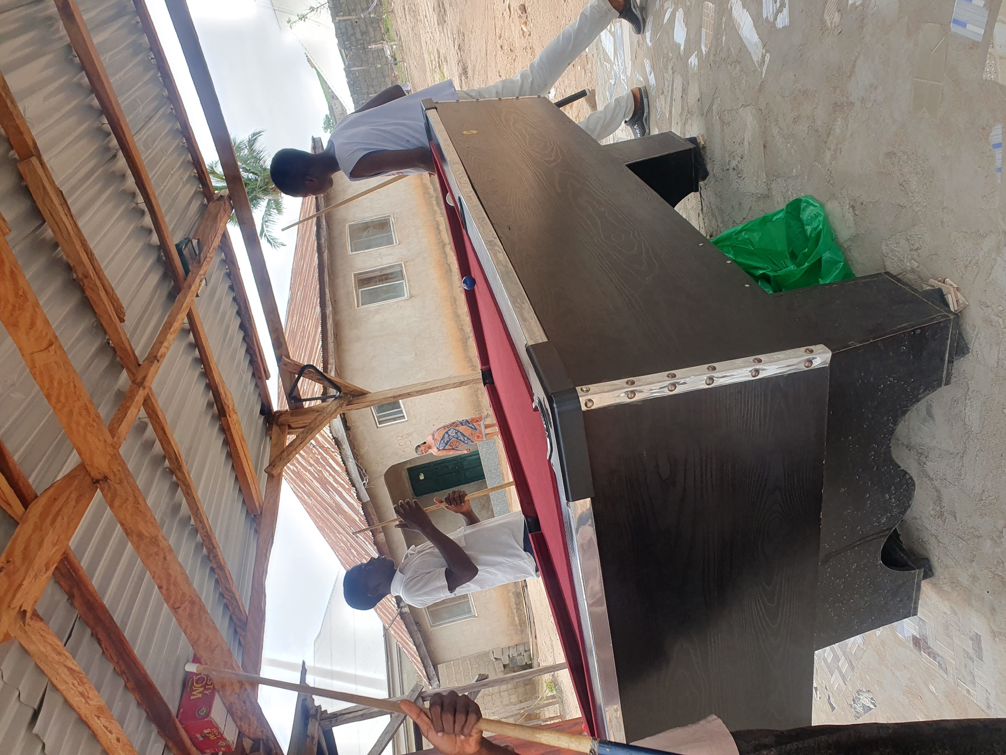 In Lokoja, Nigeria, an outdoor pool table stands on a tiled surface under a corrugated metal roof supported by wooden beams. Two young Black males, dressed casually in a white t-shirt and pants, and a white tank top and pants respectively, are actively engaged in a game of pool. One is positioned behind the table, cue in hand, seemingly lining up a shot. The other, to the left, also holds a cue, looking towards the table. A woman in a patterned dress stands in the background near a building with a reddish-brown roof, observing the game. The environment appears to be a casual outdoor setting, possibly a local hangout or residential area, with simple structures and an overcast sky. A bright green plastic bag is tucked underneath the pool table.