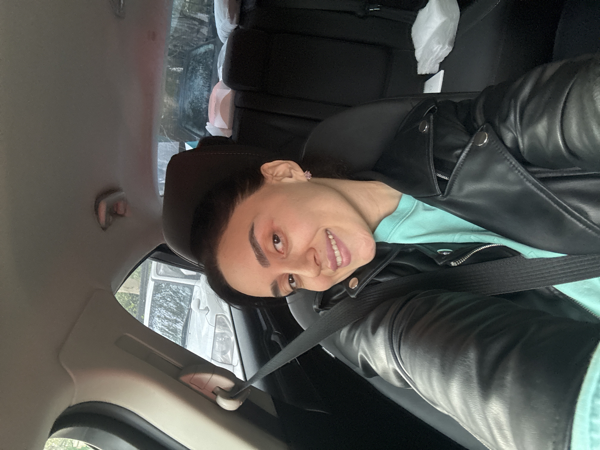 A person with dark hair, wearing a light blue shirt and a black leather jacket, is seated in the driver's seat of a car, wearing a seatbelt. The car is parked, as evidenced by a white vehicle seen through the window. The interior of the car is primarily black. The image suggests the location is Tbilisi, Georgia, based on available data and the nature of this prompt.