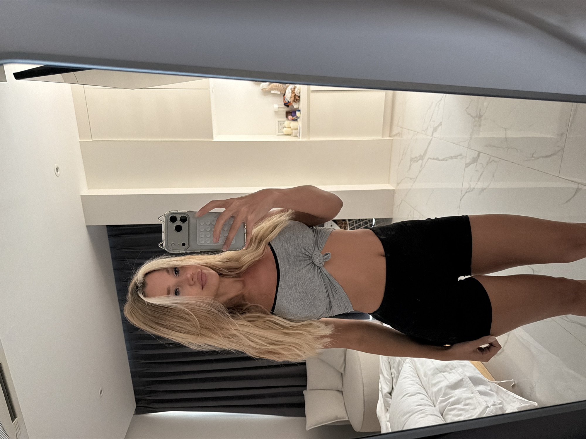 A young woman with blonde hair takes a mirror selfie in what appears to be a modern apartment. She is wearing a grey knot-front crop top and black athletic shorts. The room features a bed with white linens, a grey curtained window, and marble flooring. The background includes white cabinets and shelves with decorative items. The overall impression is of a casual, domestic setting.