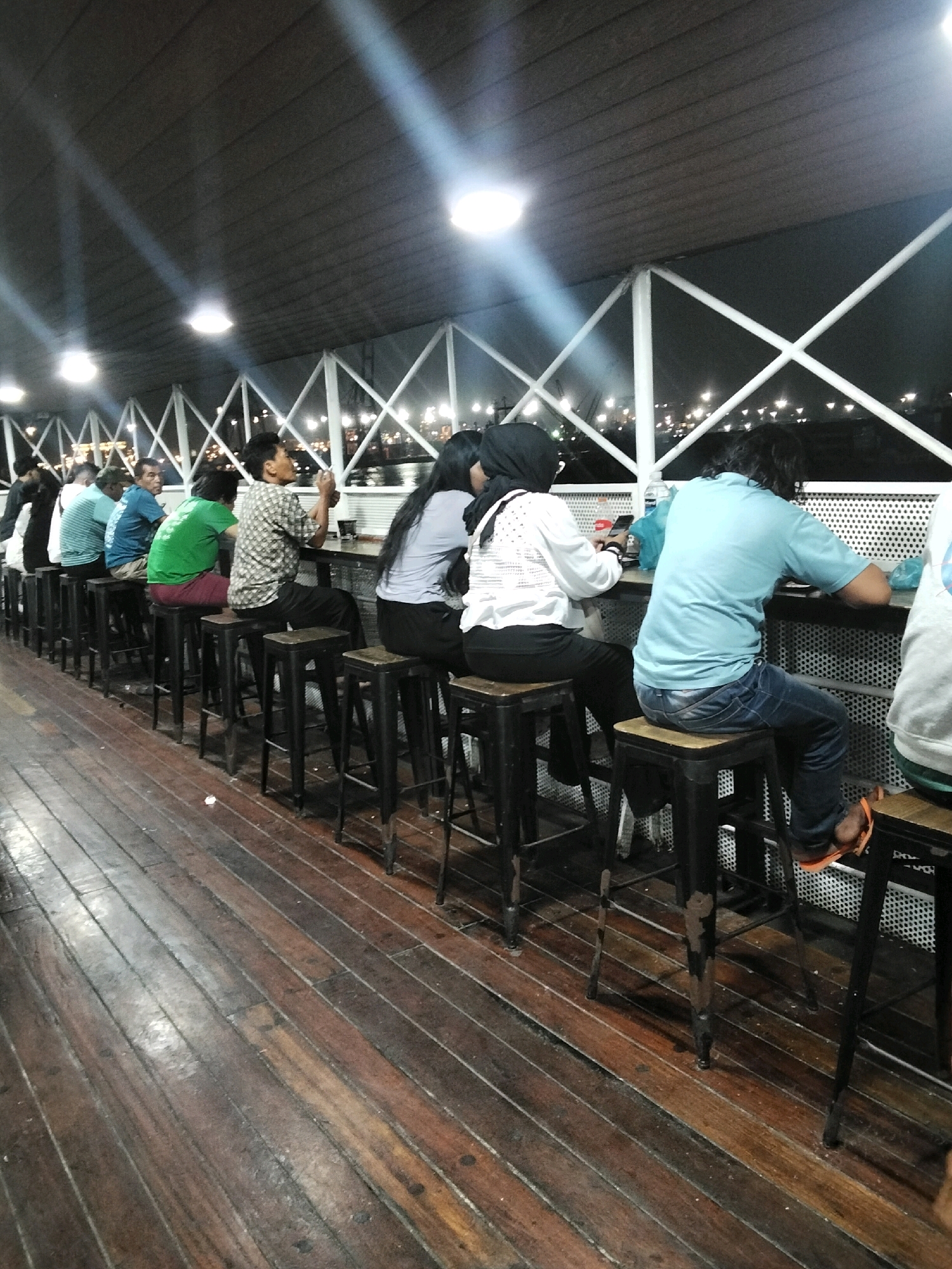 A group of people are seated on stools at a long table, which appears to be outdoors. The table is set up on a wooden deck, possibly a boardwalk. In the background, a lattice-like structure with lights is visible, suggesting a waterfront setting at night. The people are casually dressed and appear to be dining or socializing. The overall atmosphere is relaxed and informal. The location is identified as Batam City, Indonesia, suggesting a possible view of the harbor or a waterfront restaurant.