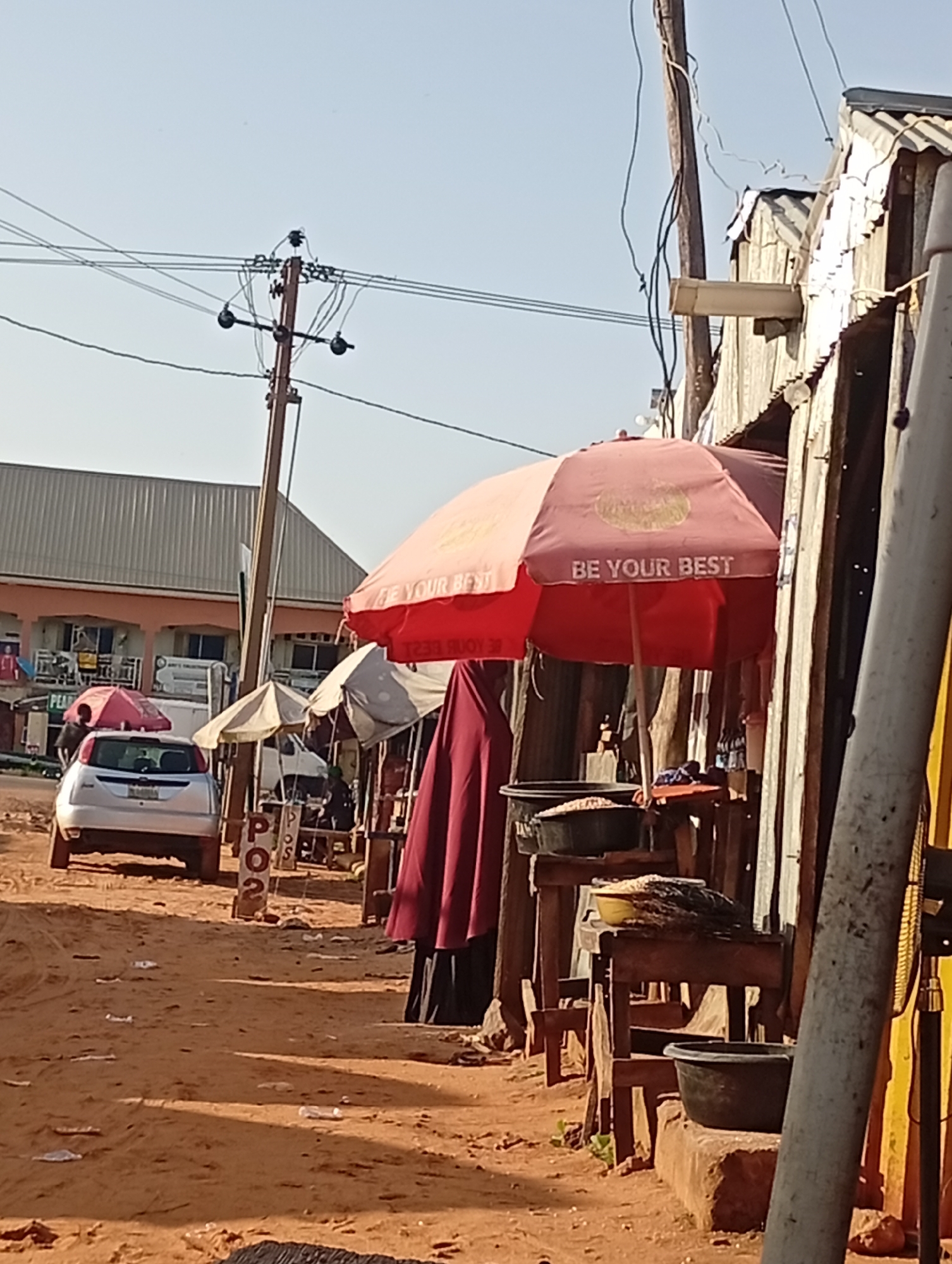 The image depicts a street scene in Tumfure, Nigeria, with a focus on a market stall area. A silver sedan is parked on a dirt road in the foreground, facing left. Behind the car, there is a white van. To the right of the car, a red umbrella is open, displaying the text "BE YOUR BEST." Several other umbrellas, some open and some closed, are visible further down the street, along with market stalls constructed from corrugated metal and wood. People are present, though mostly indistinct figures behind the stalls and umbrellas. One figure appears to be a vendor standing near the red umbrella. The ground is covered in reddish-brown dirt with scattered debris. A utility pole with electrical wires runs vertically through the scene. The environment appears to be a bustling outdoor market in a town or city setting.