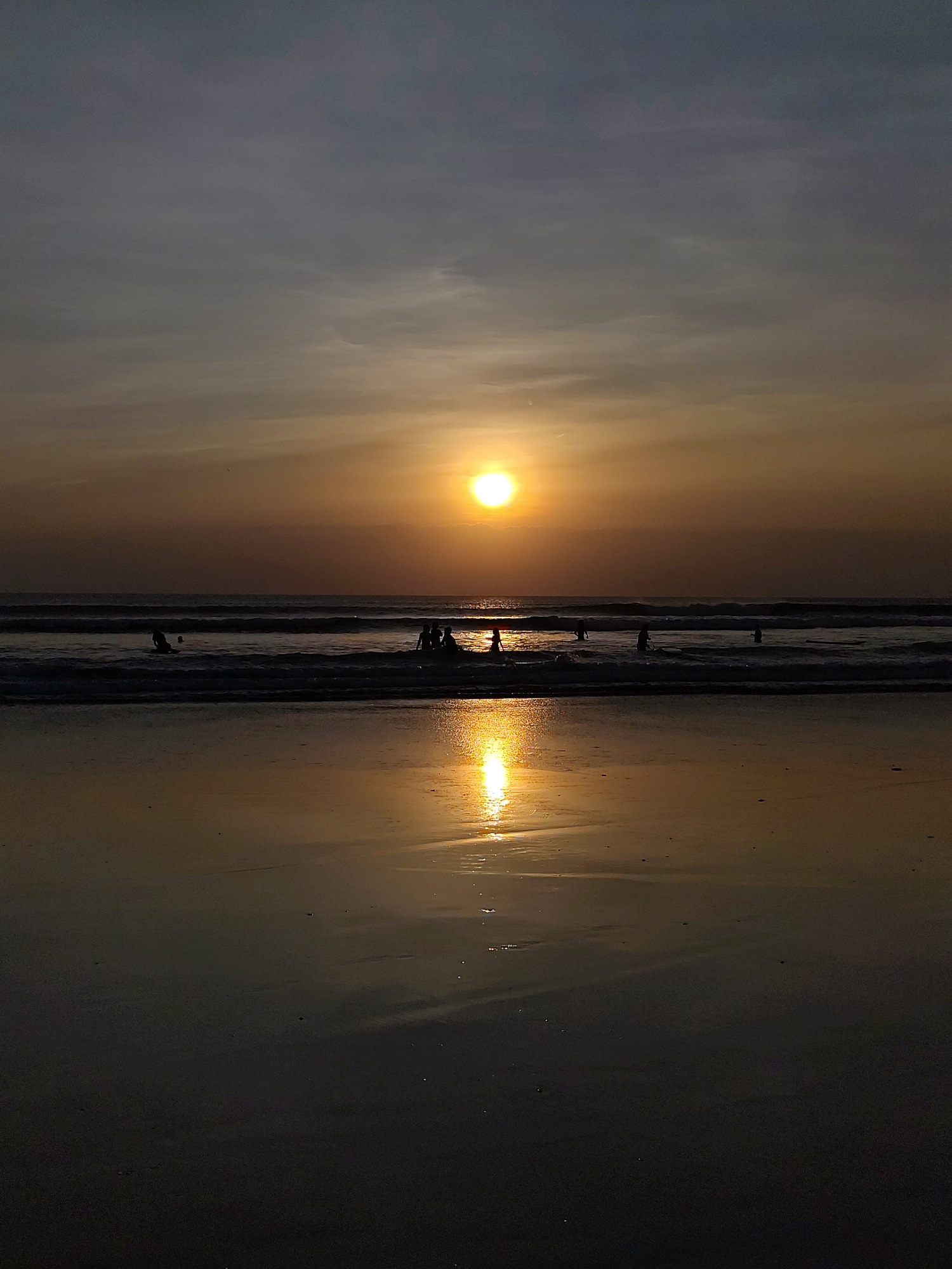 This is a vertical, full shot of a sunset at a beach. The sun is low on the horizon, partially obscured by clouds, casting a warm, golden glow across the scene. The sky transitions from a pale gray at the top to shades of orange and yellow near the sun. Waves gently roll towards a dark, wet sand beach, reflecting the light from the sun. In the water, several people are visible as dark silhouettes, some appearing to be wading or playing in the surf. The overall atmosphere is serene and peaceful, capturing a moment of natural beauty at dusk. The location is unknown.