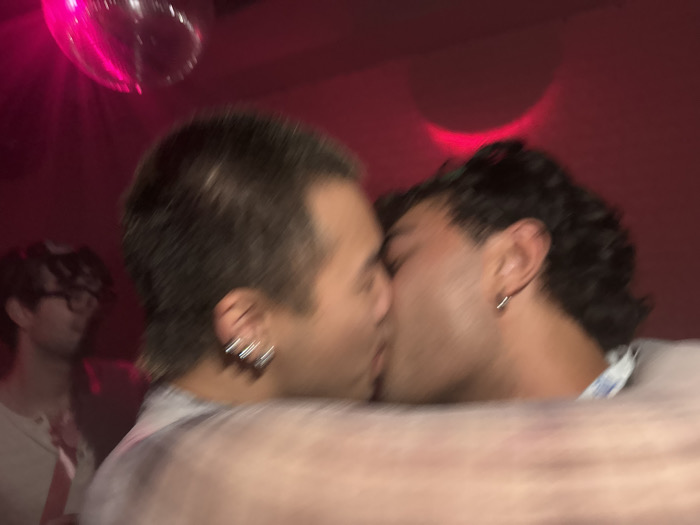 Two men are kissing in a dimly lit, red-hued room. A disco ball reflects light in the upper left corner. One man has short, dark hair with a small earring. The other has shorter hair and multiple earrings. A man wearing glasses is visible in the background. The image is blurry, obscuring finer details.