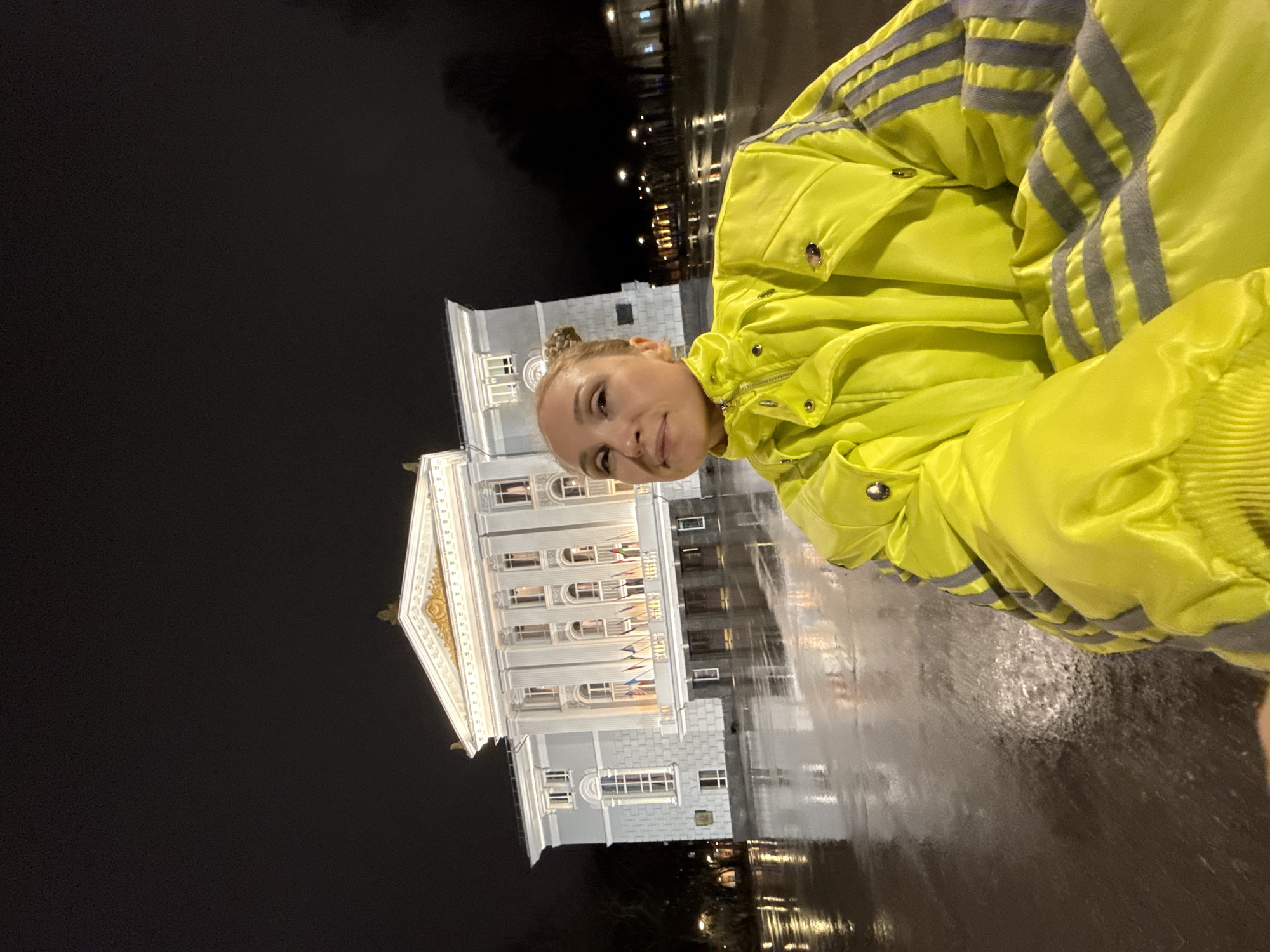 A woman wearing a yellow jacket is in front of the Perm Opera and Ballet Theatre. The photograph was taken at night, and the ground appears wet. The building is brightly lit and has a classical architectural style.
