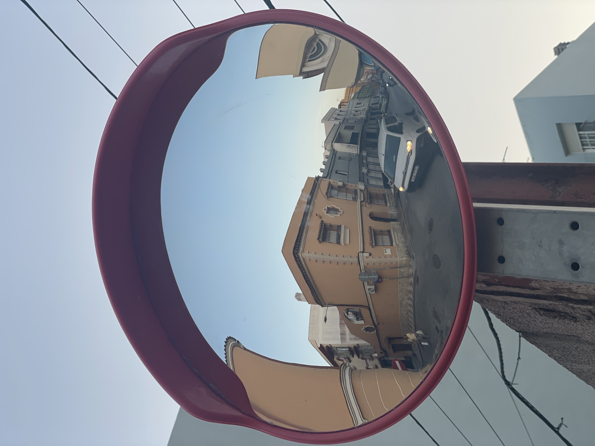 The image shows a convex mirror, likely a traffic mirror, with a red plastic frame, mounted on a metal post. The mirror reflects a street scene in La Línea de la Concepción, Spain. The reflection reveals a street lined with buildings, some of which have a light orange facade and decorative elements. A white van is visible driving on the street, with its headlights on. Further down the street, other vehicles and buildings are reflected. The sky in the reflection is a clear blue. In the foreground, outside the mirror, a rough stone surface and part of the metal post are visible, along with electrical wires.