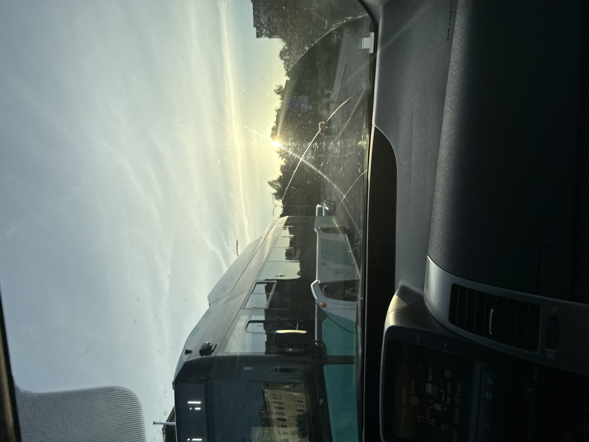 The image is taken from inside a car looking out through the windshield. The sun is low in the sky, creating a glare and lens flare on the windshield. A large bus is visible on the left side of the frame, reflecting the sky and surrounding buildings. The bus appears to be in motion. There are other vehicles and buildings visible in the background. The dashboard of the car is visible in the foreground, showing a screen displaying an "Energy monitor" with engine and battery status. The overall scene suggests a view from a vehicle traveling on a road in an urban environment, possibly during sunset. The text on the bus's display, "i il", might indicate route information or the bus company. The location is identified as Tallinn, Estonia, which suggests the bus is likely a public transport vehicle within the city.