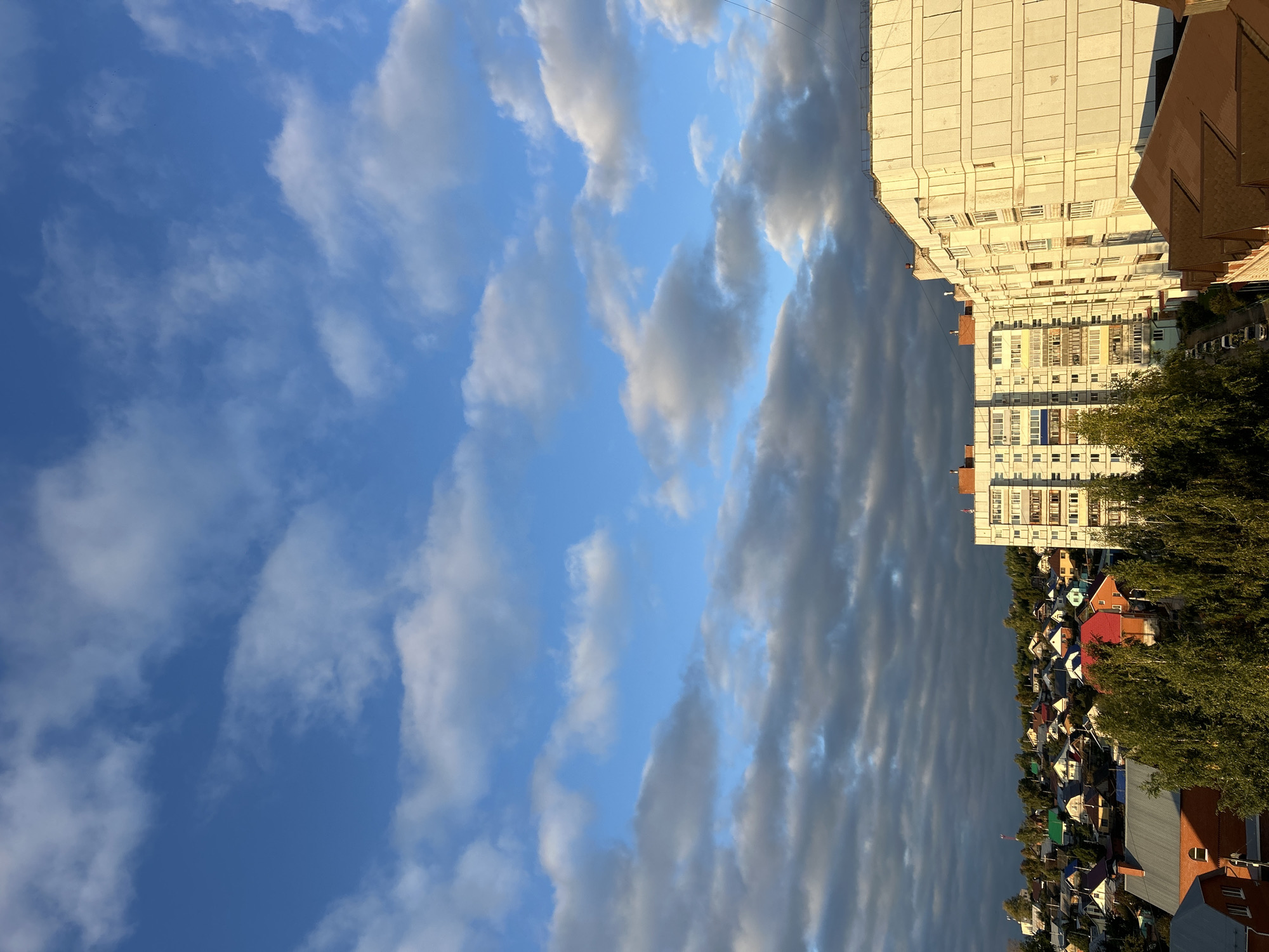 The image shows a view of Chaikovsky, Russia, on a partly cloudy day. In the foreground, there are several residential buildings, including a tall, light-colored apartment building on the right and a lower-rise apartment building in the center. A cluster of smaller houses with various colored roofs is visible in the mid-ground, nestled amongst green trees. The sky above is a vibrant blue, with scattered white and gray clouds, some of which are illuminated by the sun. The lighting suggests it might be late afternoon or early morning. There are no people visible in the image. The scene depicts a typical urban or suburban landscape with a mix of high-rise and low-rise residential structures.
