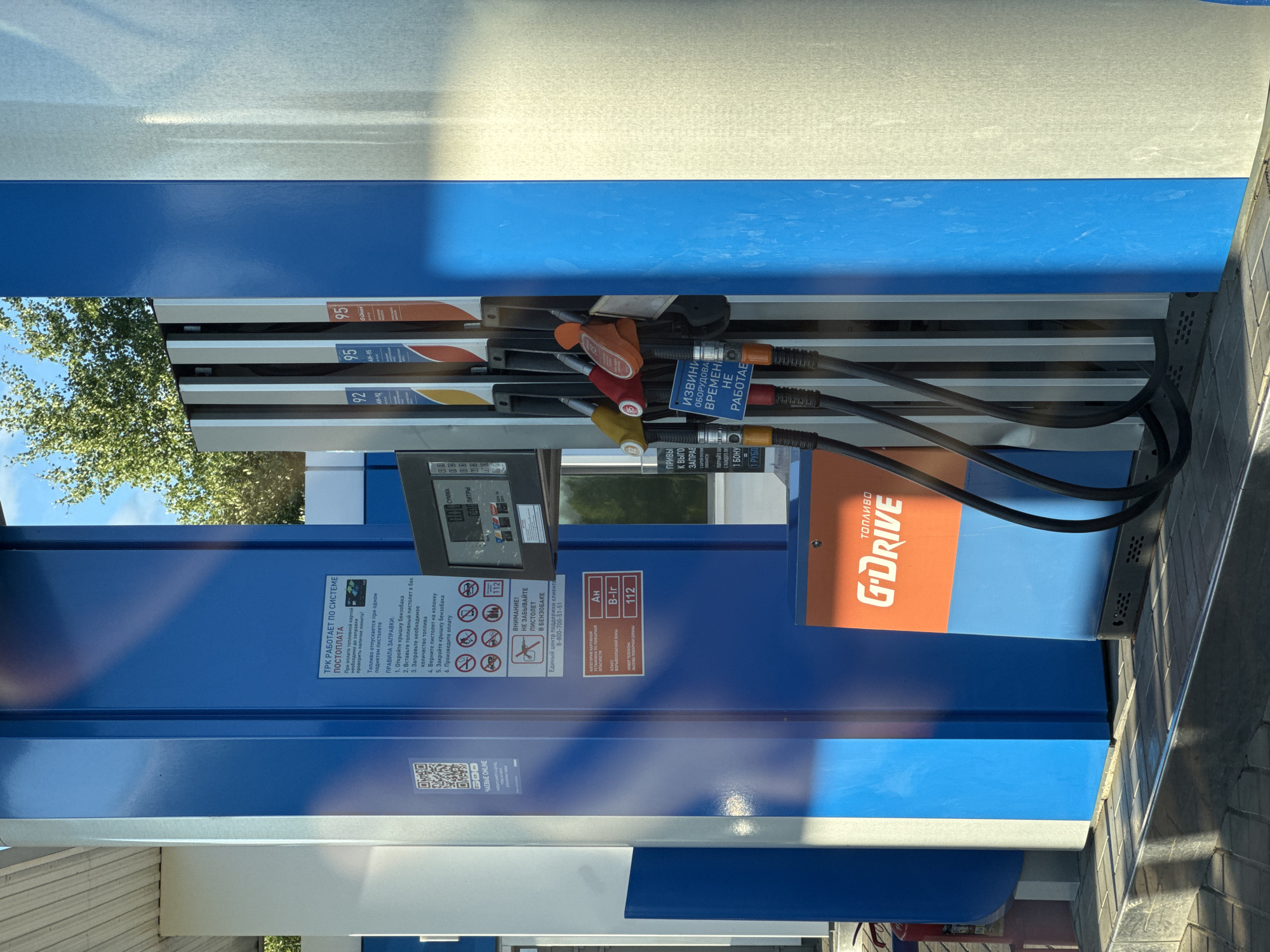This is a photograph taken at a G-Drive gas station in Pistsovo, Russia. The image focuses on a set of fuel pumps, with various gasoline nozzles visible. The pumps are predominantly blue with white and orange accents. Above the nozzles, labels indicate fuel types like "92" and "95".
On the blue panel of one of the pumps, there is a digital display showing "0.00" for "SUMMA" (sum) and "0.00" for "LITRY" (liters), suggesting no transaction is currently in progress. There is also a prominent sign with "G-DRIVE" written in bold orange letters on a blue background.
Several informational signs are visible. One instructs on the "SYSTEM POSTOPLATA" (payment system) and details the "PRAVILA ZAPRAVKI" (refueling rules), which are presented as a numbered list in Russian. Another sign in red and white warns, "Vnimanie! Ne zabyvaite pistolet v benzobake" (Attention! Do not forget the nozzle in the gas tank). There is also a contact number for a "Unified Customer Support Center."
A sign with icons and text lists hazard categories and emergency numbers, including "112" for the fire department. Text on another part of the pump reads, "Izvini oborudovano vremya ne rabotaet" (Sorry, the equipment is temporarily not working).
The scene appears to be outdoors, with a glimpse of green trees and a blue sky through the transparent cover of the fuel dispensers. The lighting suggests it is daytime, with some sunlight casting shadows and creating reflections on the surfaces. The overall impression is that of a typical, operational gas station.