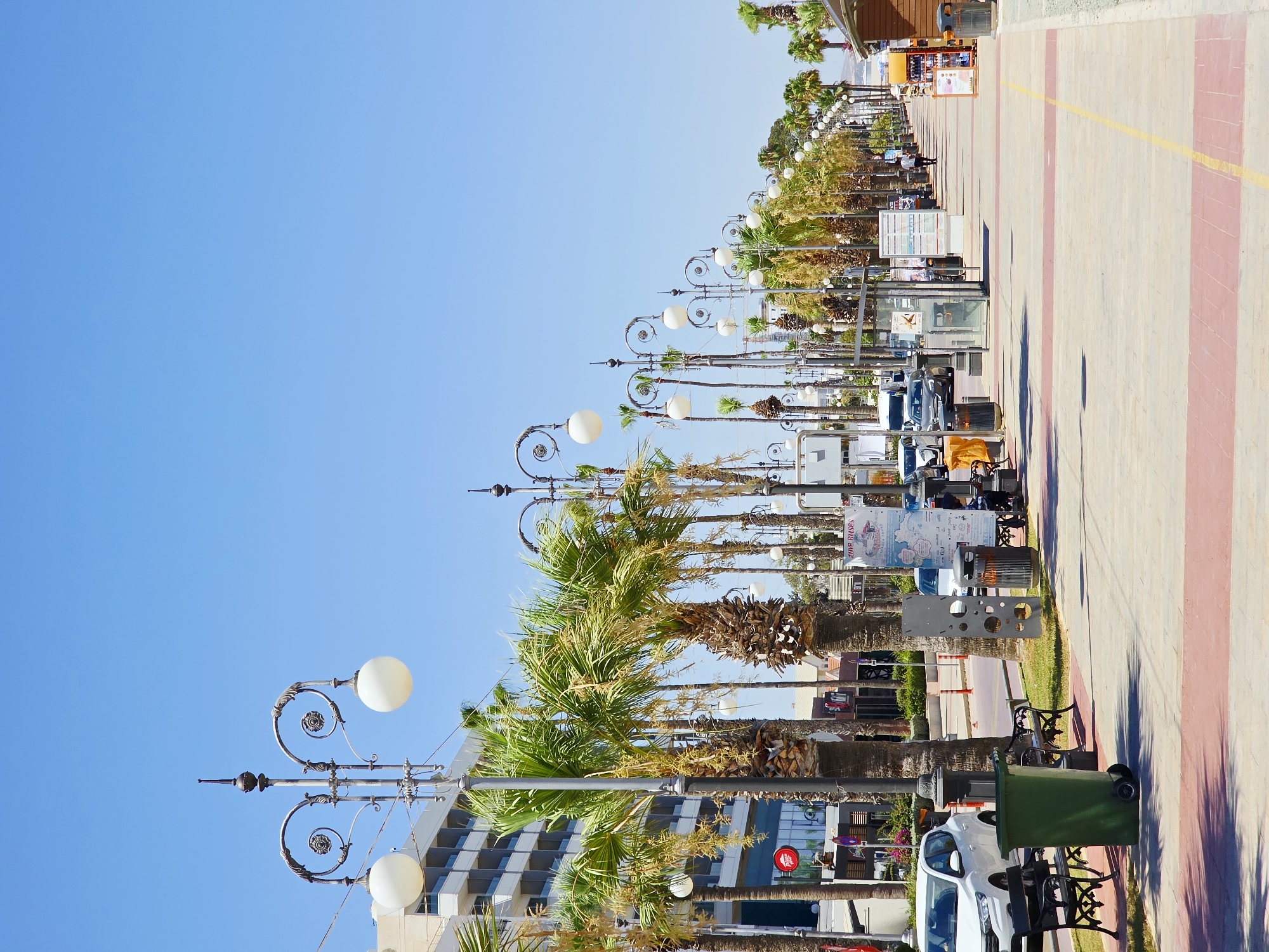 The image captures a sunny daytime scene along a pedestrian promenade in Larnaca, Cyprus, likely Finikoudes Beach promenade due to the presence of palm trees and buildings consistent with a coastal resort. The promenade is paved with a light-colored stone material, marked with red and yellow stripes. A row of ornate lampposts with white globe lights lines the left side of the promenade, interspersed with numerous tall palm trees.
On the left side of the promenade, behind the lampposts and palm trees, is a modern, multi-story hotel building with a light-colored facade. Below the hotel are various shops and establishments, one with a red circular logo that appears to be Pizza Hut. A white car is parked in front of the hotel, alongside a green trash bin and two black park benches.
The promenade extends into the distance, where more palm trees and lampposts continue. Several vehicles, including cars and a white van, are visible on the road running parallel to the promenade. People are seen walking along the promenade in the distance, suggesting a moderately active public space. Some information kiosks and bus stop shelters are also present along the promenade.
The sky is clear and bright blue, indicating good weather. The overall impression is one of a vibrant, well-maintained tourist area typical of a Mediterranean coastal city.