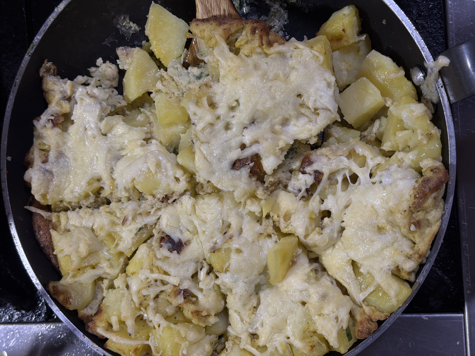 A top-down shot shows a frying pan filled with an omelet containing potato chunks and topped with melted cheese. The frying pan is black with a metal handle. The food appears to be cooked and ready to eat. The setting appears to be a kitchen, with a stovetop or countertop visible around the frying pan. It is not possible to determine if the image was taken in Yerevan, Armenia. There are no people visible in the image.