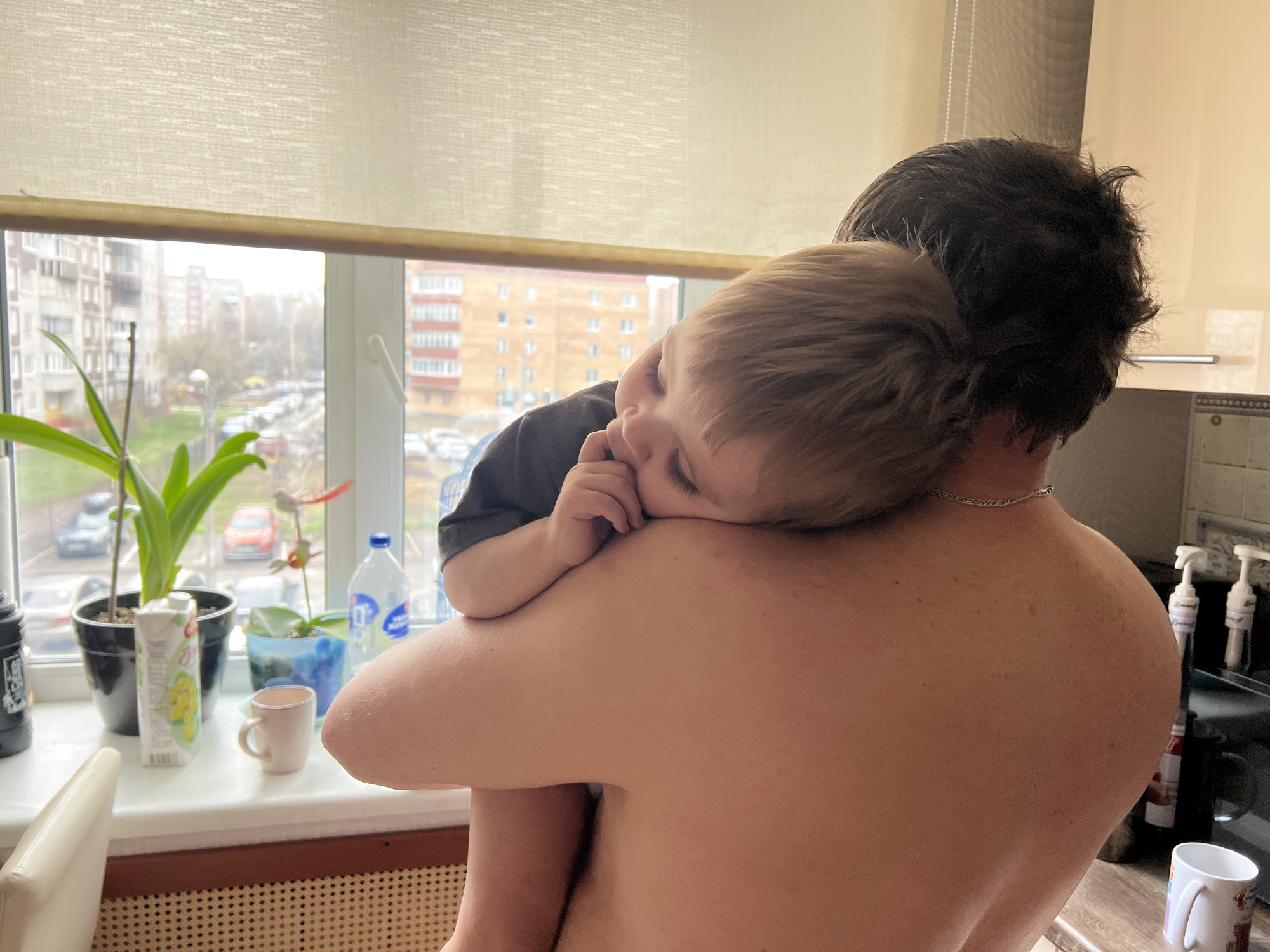 A shirtless adult is holding a child, who is wearing a dark gray t-shirt, on their shoulder. The child is sleepy and has their eyes closed. The location appears to be indoors near a window. A table or counter is visible near the window, upon which are several plants, a juice carton, and a water bottle. Outside the window, apartment buildings and cars suggest an urban environment. Based on the architectural style of the buildings visible in the window, the image may have been taken in Perm, Russia. A backsplash is visible on the right, as well as items on the counter.
