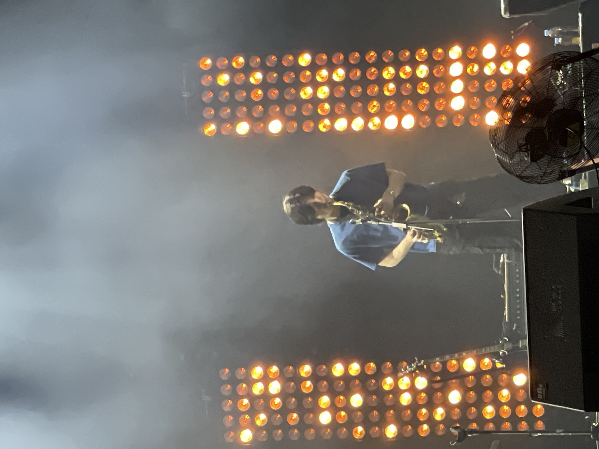 A musician plays a saxophone on stage, illuminated by two sets of grid-patterned stage lights with orange bulbs. A black fan is visible on the right side of the stage. The stage is filled with smoke or haze. The image was taken indoors, likely in Salembaran, Indonesia.