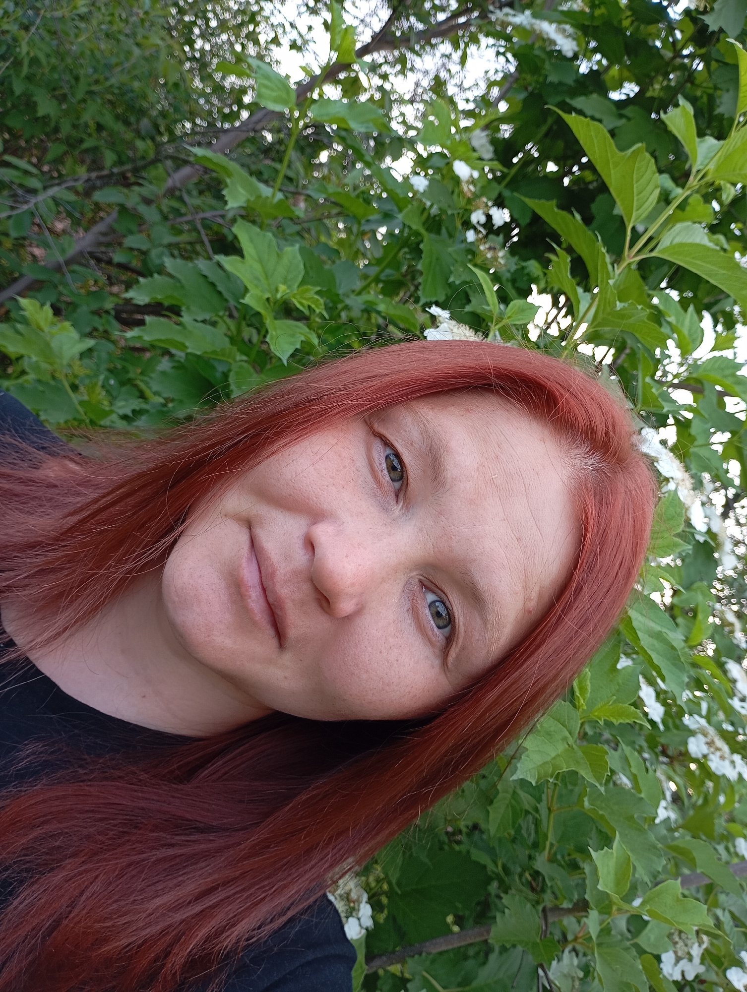 A selfie of a woman with red hair. She is wearing a black shirt, and has light colored eyes. She is standing in front of a bush with green leaves and white flowers. The location is Фоки, Russia.