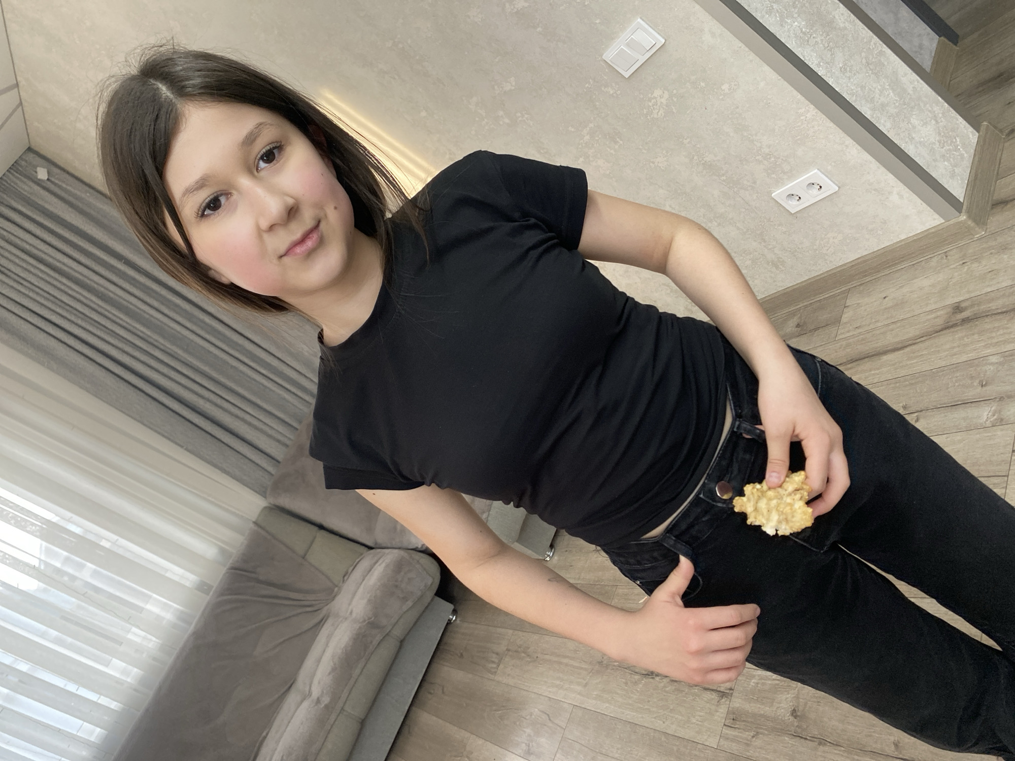 A young person, likely a teenager or young adult, is depicted in an indoor setting in Izhevsk, Russia. She stands at an angle, looking directly towards the camera with a calm, slightly neutral expression. She has dark, shoulder-length hair and is dressed in a plain black short-sleeved t-shirt and black jeans. Her left hand is positioned near her hip, holding a piece of a light-colored, irregular-shaped snack, resembling a granola cluster or oatmeal cookie.
The background shows a contemporary room featuring light wooden floorboards and a light grey or beige textured wall/ceiling. To the left, a portion of a grey upholstered sofa is visible, with light-colored, possibly sheer, curtains or blinds covering a window behind it. On the upper part of the wall or ceiling, white rectangular light switches and a white double European-style electrical outlet are present. The image is taken from a slightly low and tilted perspective, emphasizing the subject and the interior design elements. The overall scene conveys a casual, everyday moment in a modern home.