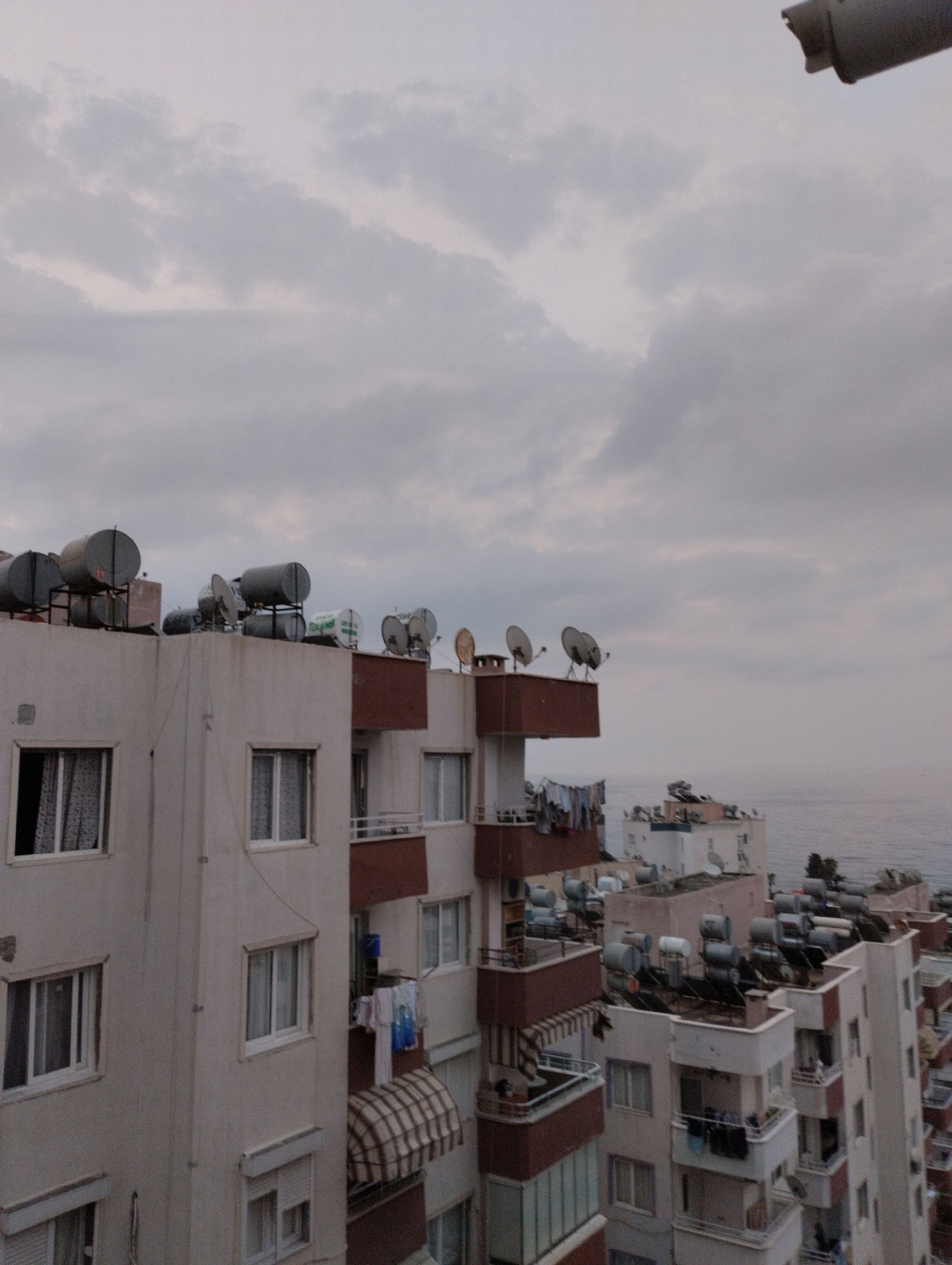 The image captures a view of apartment buildings in Mezitli, Turkey, under a cloudy sky. The buildings are multi-story structures with many windows and balconies. On the rooftops of several buildings, numerous solar water heaters are visible, along with satellite dishes. Laundry is hanging on clotheslines on some balconies. The buildings are situated close to the sea, which is visible in the background. The overall atmosphere is overcast and somewhat muted in color.