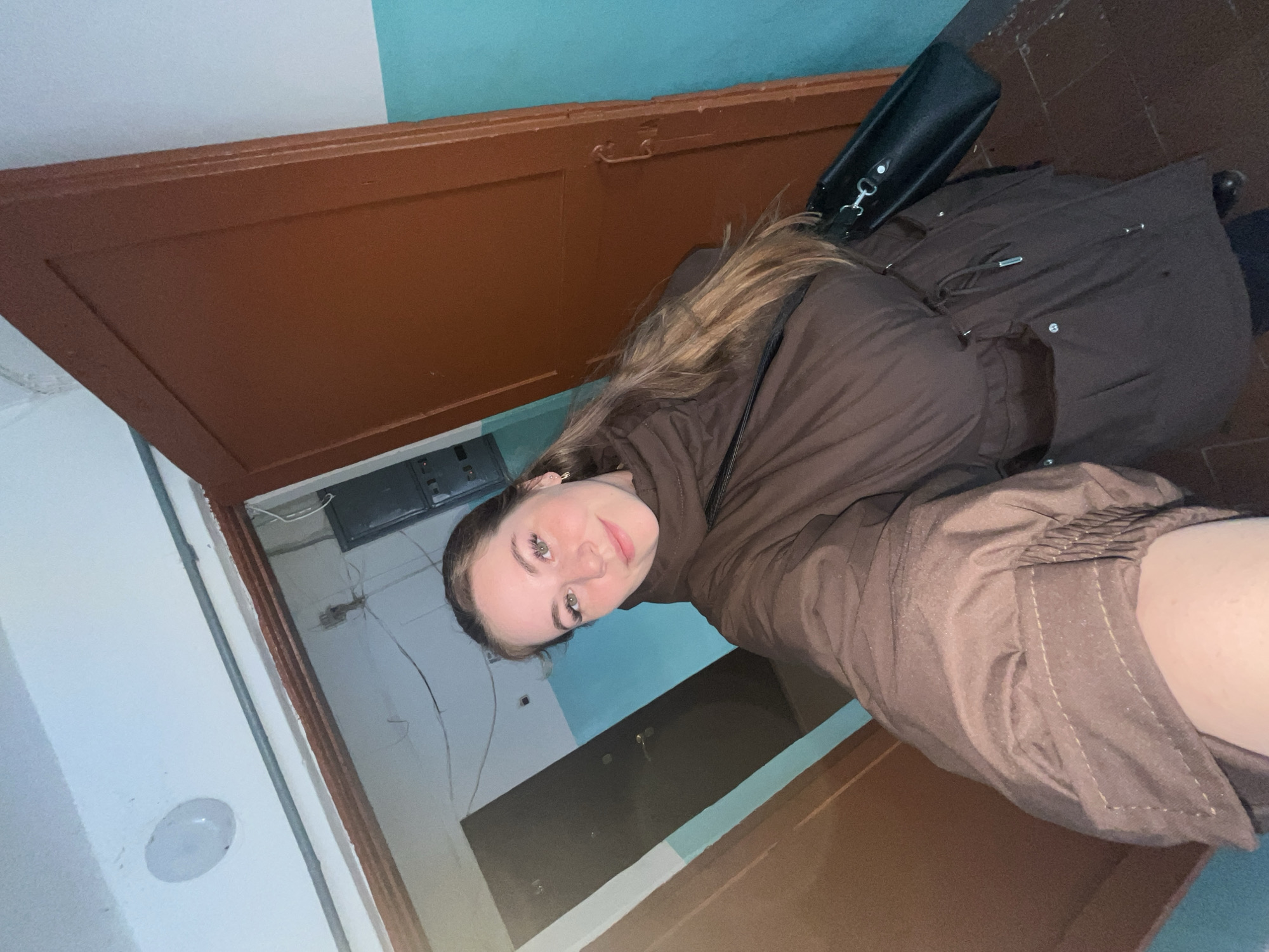 A young woman is depicted in a selfie-style photograph taken indoors, likely in a hallway or entryway of a building in Chaikovsky, Russia. She has long brown hair, green eyes, and is looking directly at the camera with a slight, pleasant smile. She is wearing a brown puffer-style coat and has a black shoulder bag visible on her right side.
The background features a two-toned wall, with the upper section painted a light blue and the lower section white. A brown wooden door is prominent on the left side of the frame, and another brown door or doorframe is visible in the lower right. Exposed electrical wires and a gray electrical box are visible on the wall behind the woman. A white, circular light fixture is mounted on the white wall section. The flooring appears dark and utilitarian.
The woman is the central subject, engaged in the activity of taking a self-portrait. The indoor setting, with its artificial lighting and visible infrastructure elements, suggests a functional area rather than a decorative one. There is no discernible text within the image.