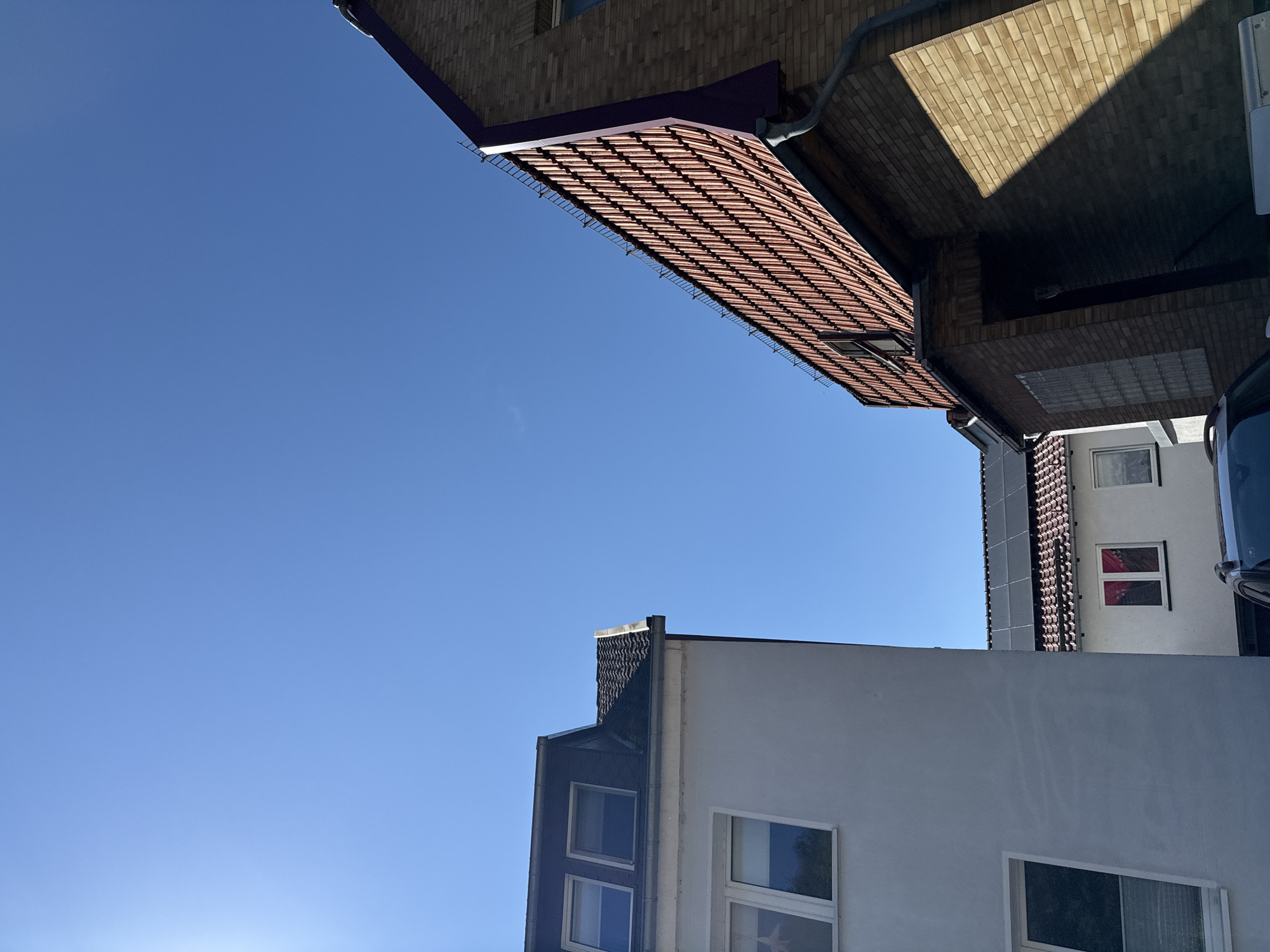 The image depicts a street scene in Bad Sachsa, Germany, on a clear, sunny day. Two buildings are prominently featured. On the left, a white building with a mansard roof is visible, featuring several windows. To the right, a brick building with a steeply pitched, tiled roof dominates the frame. This building has a darker, purplish trim along the edge of the roof and a visible gutter system. Solar panels are installed on the roof of a smaller building situated behind the brick structure. The sky is a vibrant, cloudless blue, indicating fair weather. The perspective suggests the photo was taken from street level, looking upwards at the buildings.