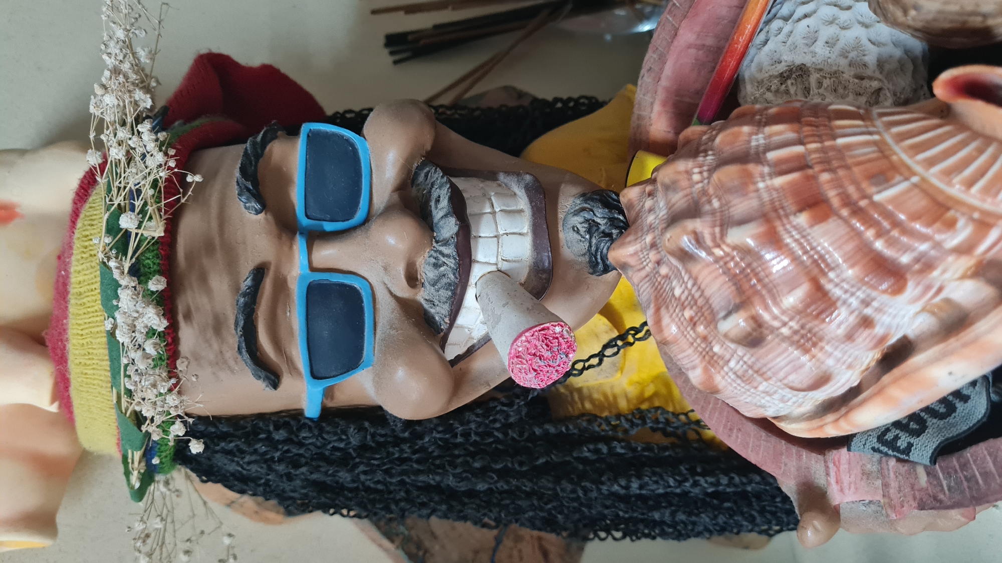 A close-up shot, taken indoors, presents a horizontally oriented, sculpted head wearing blue sunglasses and a multi-colored hat with dried white flowers. The sculpture has dark, textured hair. A cigarette with a pink tip is positioned in the sculpture's mouth. The sculpture is adjacent to a large seashell. The location is Lachish Regional Council, Israel.