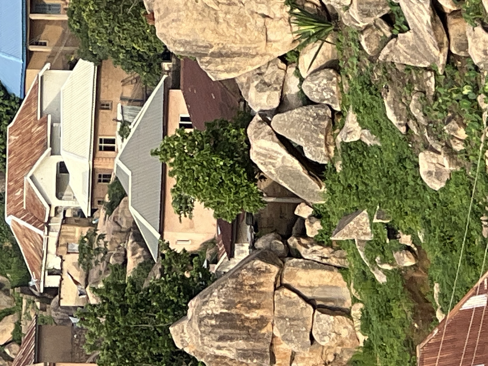 The image depicts a rugged, hilly landscape in Jos, Nigeria, characterized by a dense cluster of buildings integrated with large rock formations. The architecture is varied, with many structures featuring corrugated iron roofs in shades of brown and grey, alongside some with lighter colored metal roofs. Some houses appear to be constructed from concrete and cement, with painted walls in earthy tones. Vegetation, primarily lush green shrubs and trees, is abundant, growing amidst the rocks and around the buildings, suggesting a natural, somewhat untamed environment. The scene conveys a sense of a community built into and around a rocky terrain, with the buildings following the contours of the land. There are no people or animals visible in the frame.