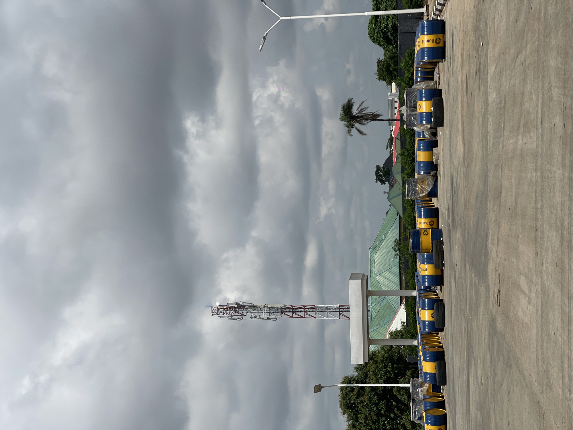 In Abuja, Nigeria, a cloudy sky looms over an outdoor storage area filled with numerous blue and yellow barrels. The barrels appear to be of uniform size and are arranged in multiple rows across a paved surface. Some barrels are covered with transparent plastic sheeting. In the background, a tall communication tower stands prominently, its metallic structure visible against the overcast sky. Adjacent to the tower is a white structure with pillars, possibly a shelter or canopy. Further in the distance, buildings with green roofs are visible, along with trees and a palm tree. A street light stands to the left of the frame, and another to the right. The scene suggests a commercial or industrial setting.