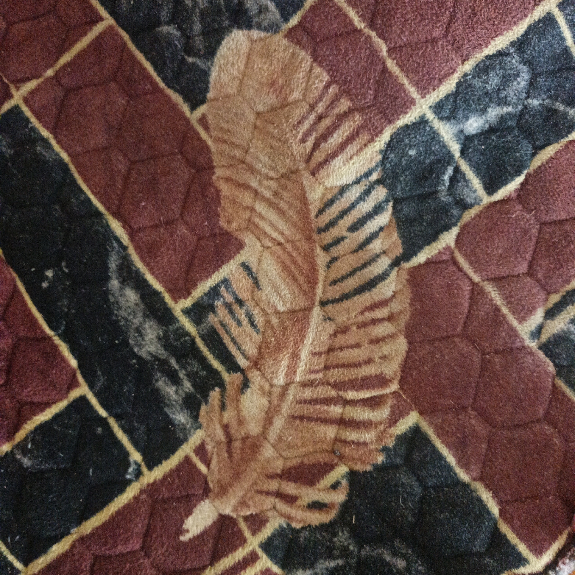 This is a close-up, top-down, detailed view of a woven fabric with a pattern. The pattern consists of geometric shapes, primarily diamonds and hexagons, formed by a grid of golden yellow lines against a background of maroon and dark grey/black. Superimposed on this geometric pattern is an illustration of a light brown feather. The feather is detailed with darker brown striations, suggesting quill and barbs. The fabric appears textured and possibly padded, with a soft, slightly plush surface. The lighting is even, casting no significant shadows, and the image is sharp, allowing for observation of the fabric's weave and texture. There are no people, animals, or any recognizable elements of the city of Sungai Rasau, Indonesia, present in the image.