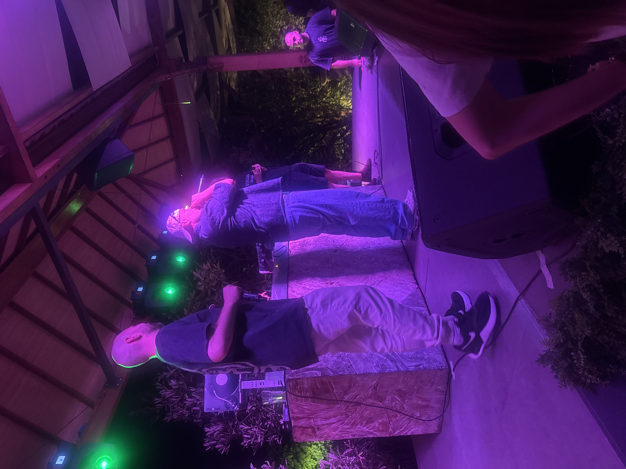 The image shows an outdoor performance stage at night, illuminated by purple and green lights. On stage, there are three men. The man on the left, with a shaved head, is wearing a black t-shirt and light-colored pants, standing near a DJ booth with a turntable. The man in the center, wearing a cap and denim shirt, is holding a microphone and appears to be rapping. To the right, another man in a black t-shirt stands near a speaker. In the background, lush greenery is visible. The stage is covered by a wooden roof structure. The image is captured from a low angle, focusing on the performers and the stage area. The scene appears to be from an event in Varna, Bulgaria, possibly at the "Sea Station" venue given the architectural style and outdoor setting.