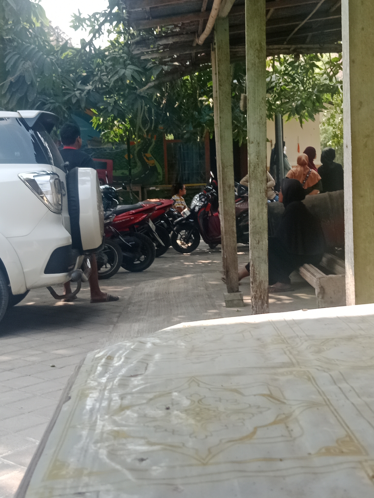 In Srikaton, Indonesia, a white SUV is parked on a paved area next to a covered structure with wooden posts and a corrugated metal roof. Several motorcycles, including a red one and a black one, are parked behind the SUV. In the background, a group of people, some wearing headscarves, are gathered under the covered structure, seated on a bench. The environment appears to be an outdoor seating area with trees and foliage visible. A table with a patterned surface is in the foreground.