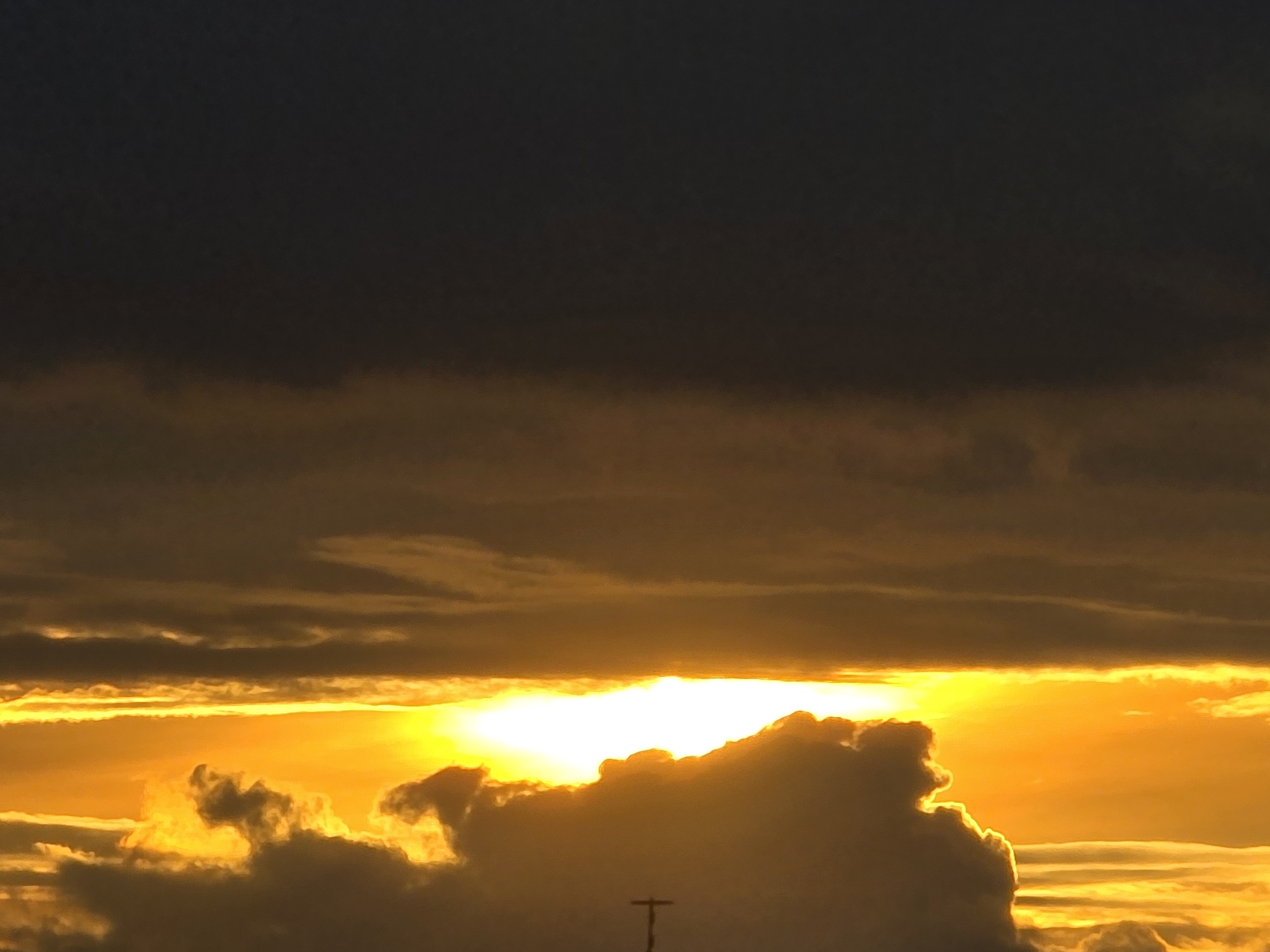The image shows a sunset over Vilnius, Lithuania. The sky is filled with dramatic clouds, with the sun peeking through a gap, casting a golden glow. The lower part of the sky is a vibrant orange and yellow, while the upper part is a darker, muted gray. In the foreground, a silhouette of a cross-shaped structure is visible against the bright sky. The scene suggests a peaceful, natural phenomenon occurring within an urban environment, hinting at the presence of a church or other religious building with a prominent cross.