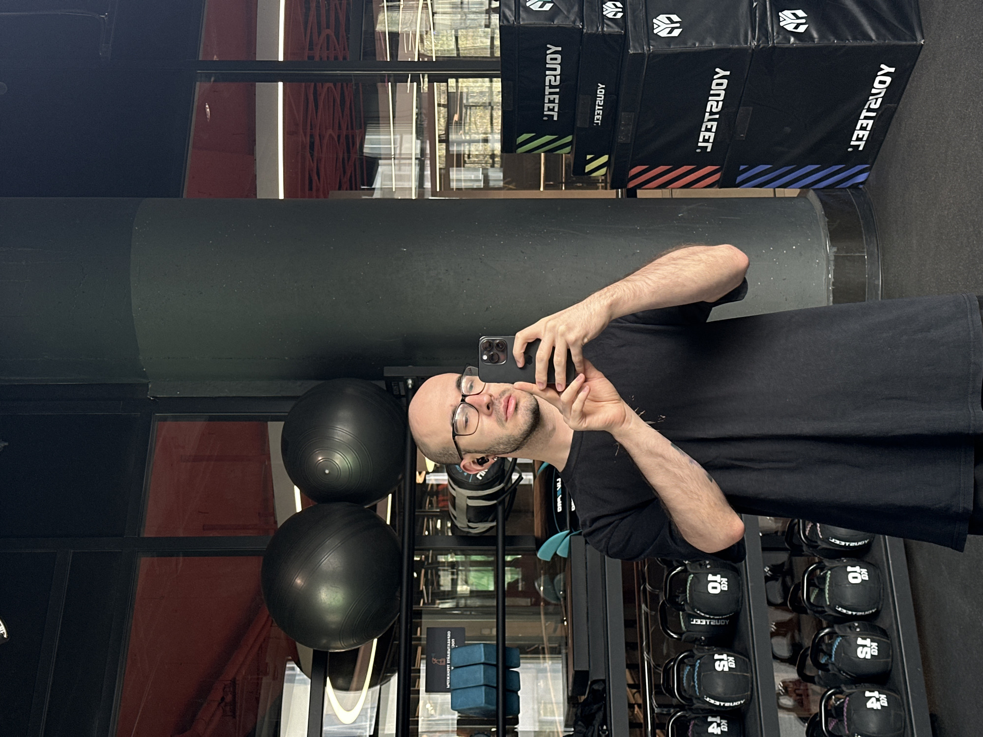 A man stands in a gym, holding a smartphone and taking a selfie. He has short hair, a beard, and wears glasses. He is wearing a black t-shirt. Behind him, there is exercise equipment including medicine balls, weights, and plyometric boxes. The gym appears to be in a building with large windows, but the exact location in Moscow, Russia cannot be determined from the available visual information.